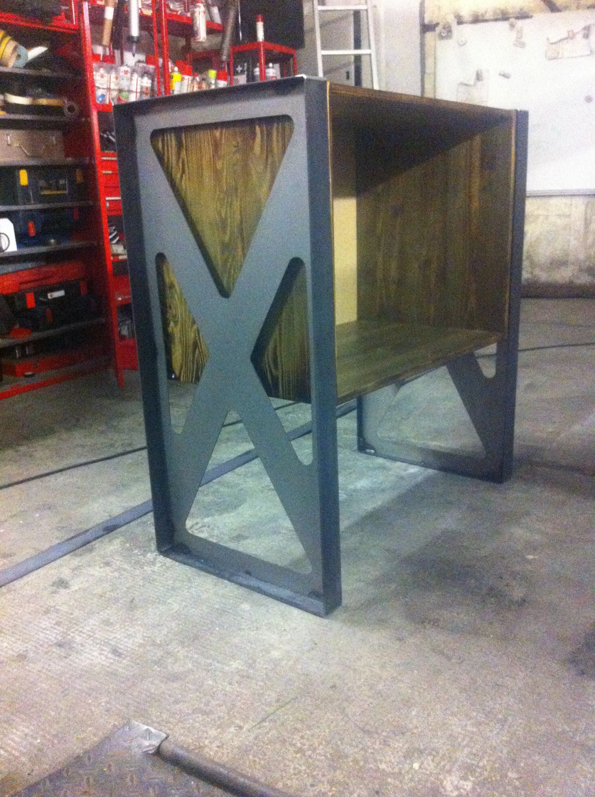 A custom desk with a wood finish and metal frame featuring large cutouts of music note symbols, set in a workshop with tools and red storage shelves in the background.