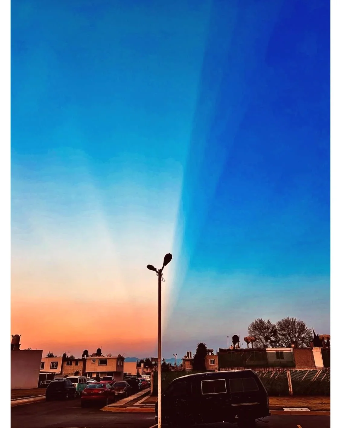 A parking lot at sunset with cars, a lamp post, and trees in the background, under a sky with a sharp color divide between warm sunset and cool blue twilight.