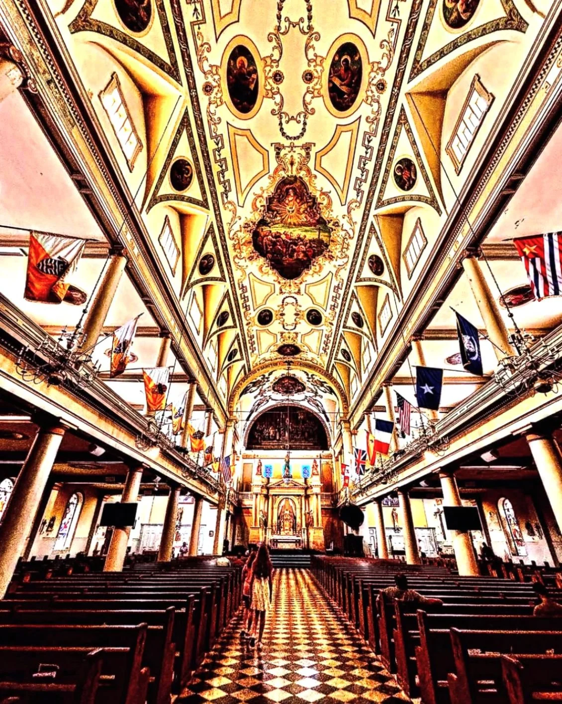 Interior of a church with ornate ceiling murals, flags hanging from the balcony, wooden pews, and an altar at the front.