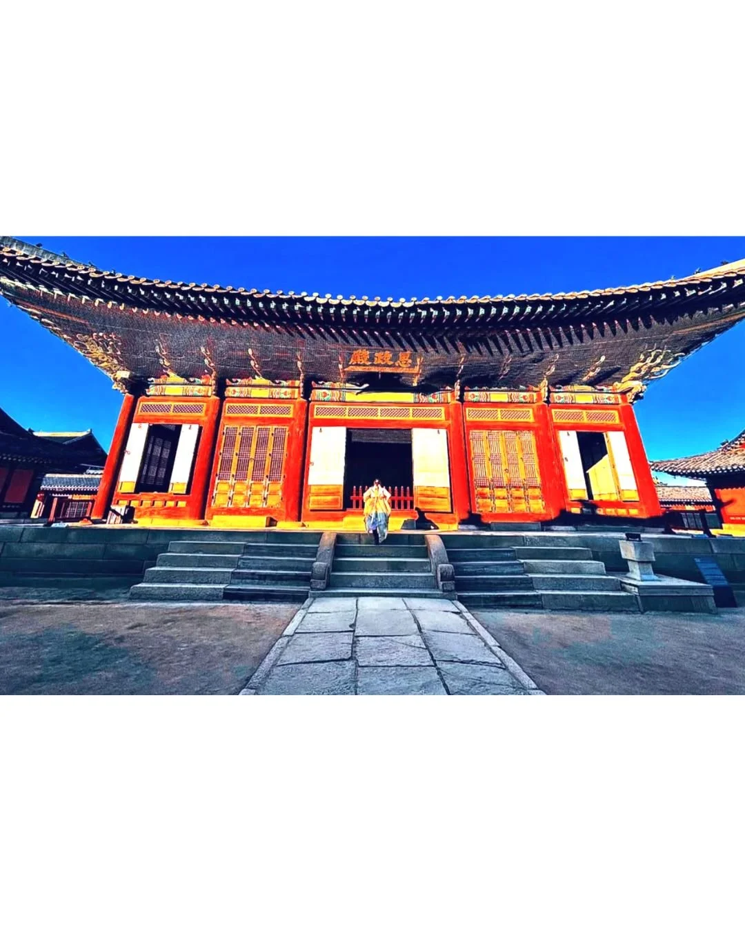 Traditional Korean palace with wooden architecture, red and yellow doors, and a wide staircase leading to the entrance, under a bright blue sky.