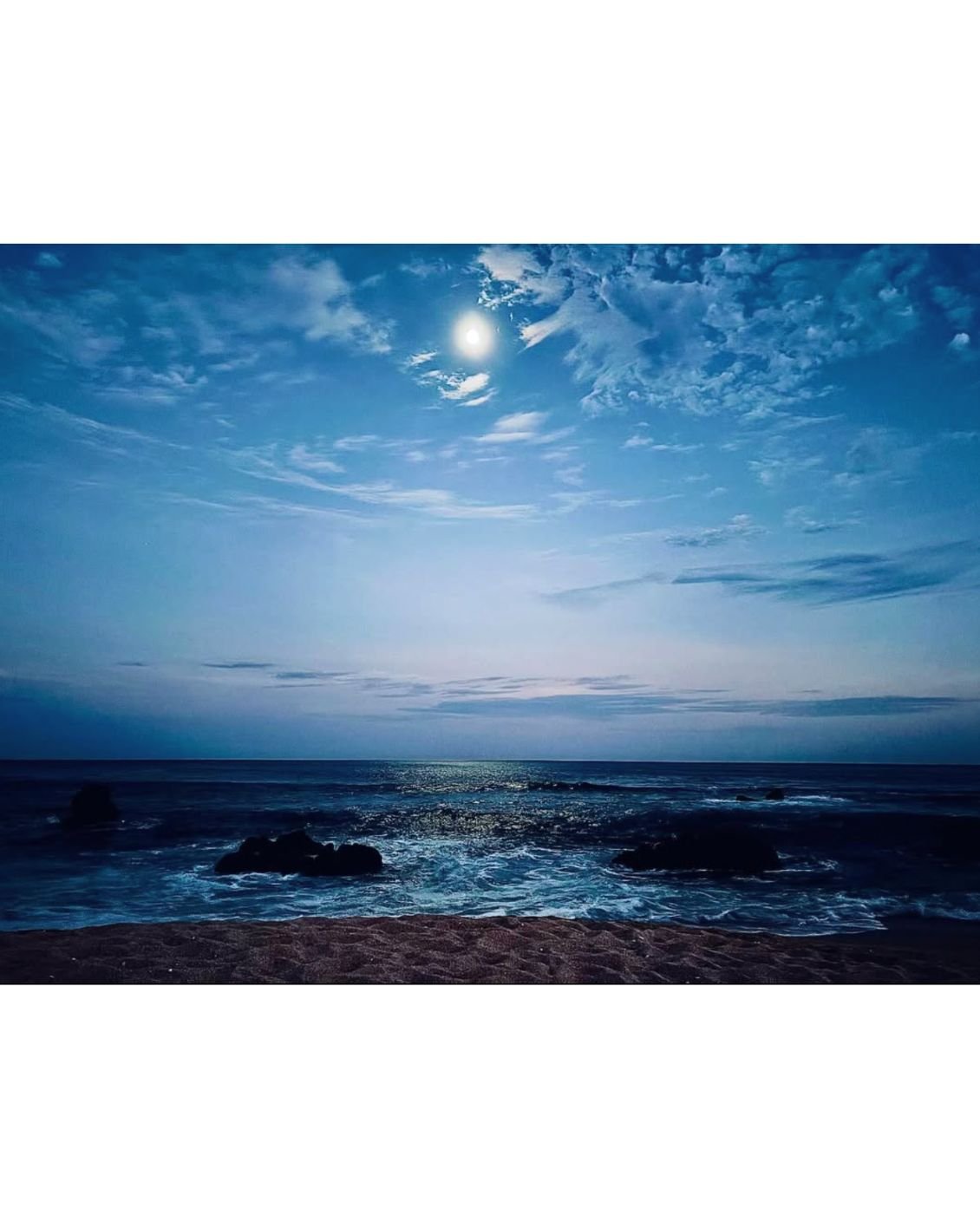 A beach scene under a moonlit sky, with clouds illuminating the night and waves crashing against rocks along the shore.