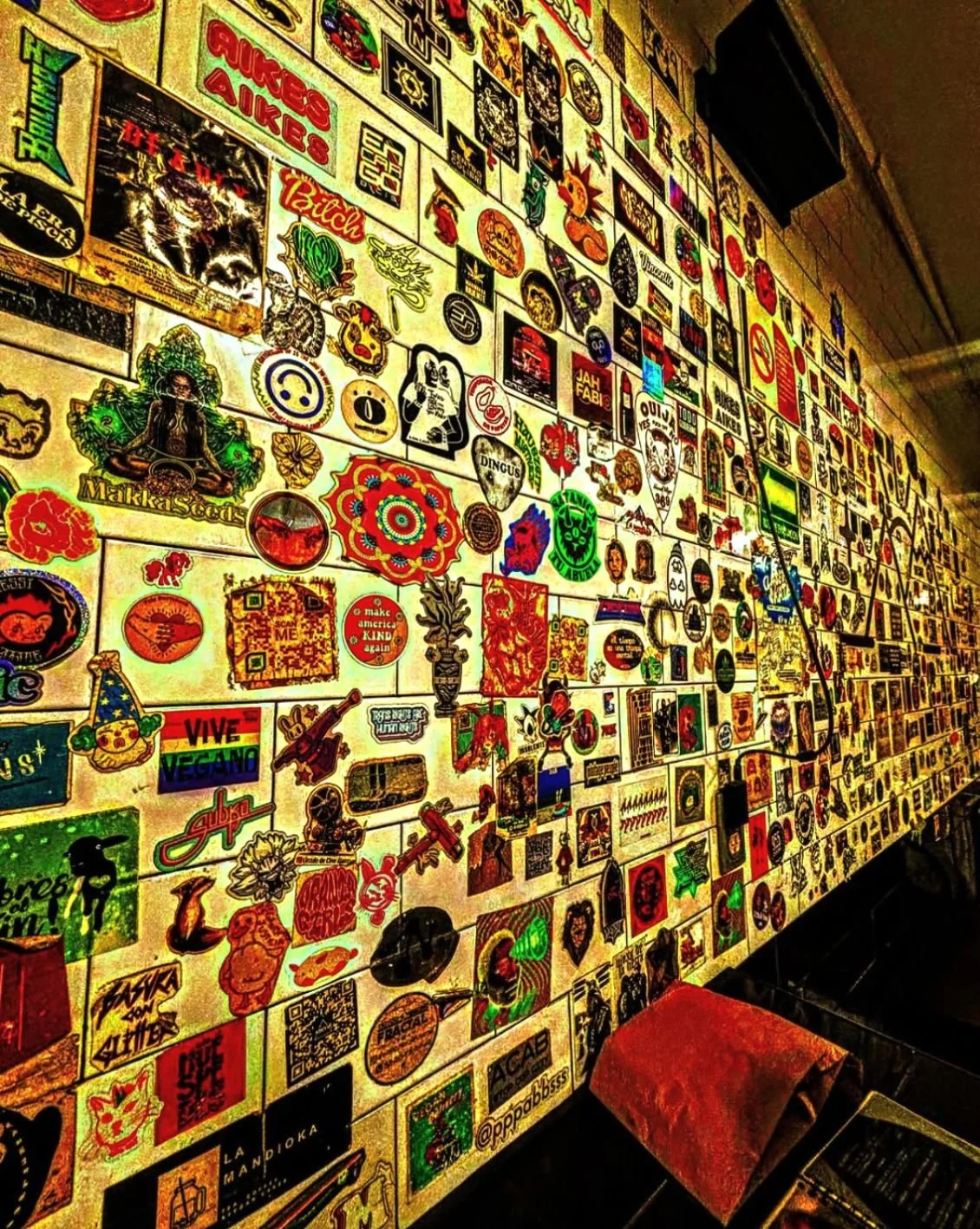 A wall covered with numerous colorful stickers and decals of various shapes and designs, including logos, characters, symbols, and text, with a cloth-covered bench in front.