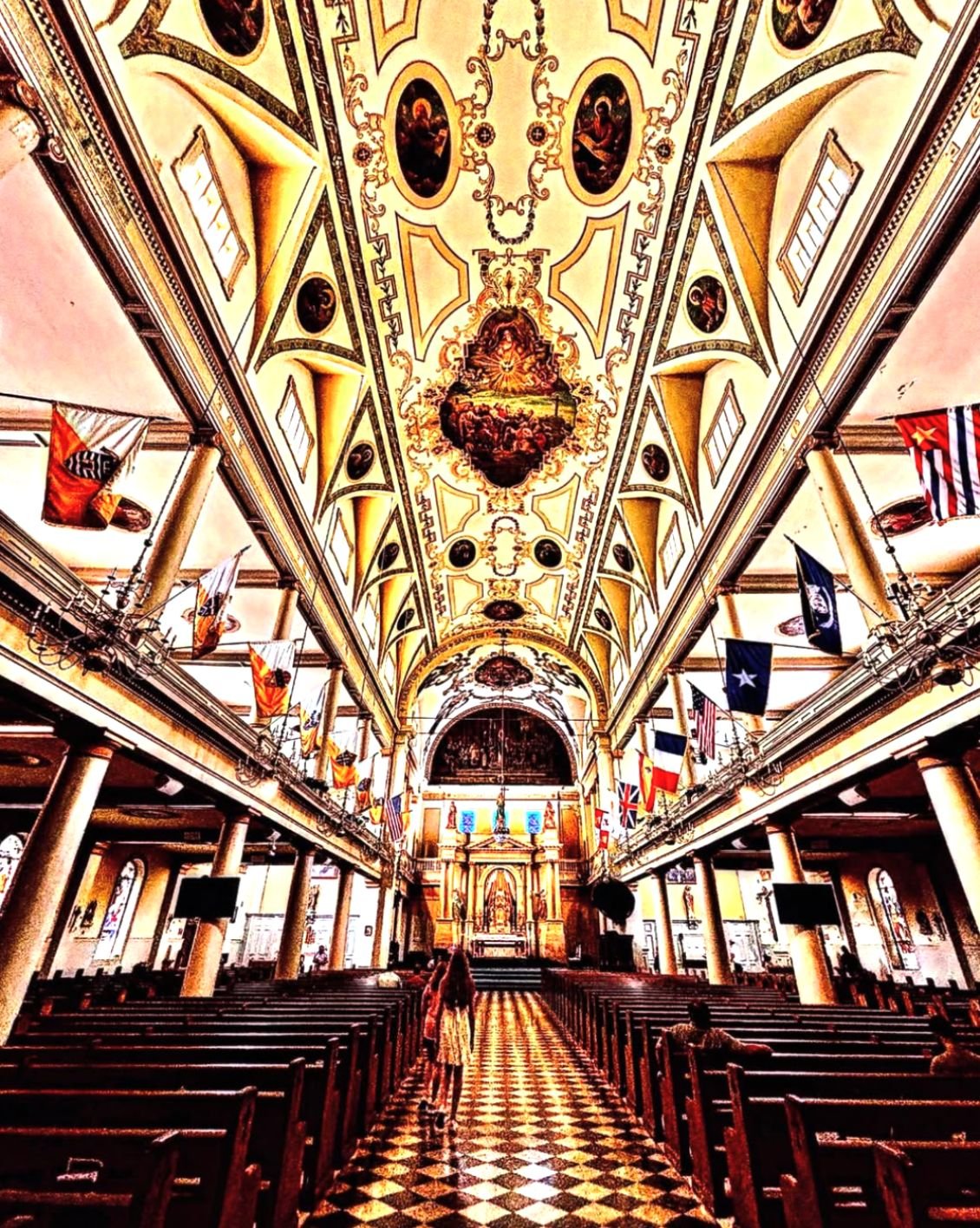 Interior of a church with ornate ceiling, religious paintings, flags hanging from the balcony, wooden pews, and checkered floor.