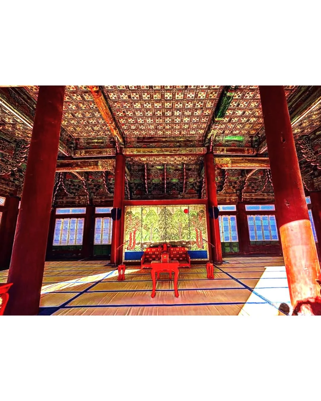 Interior of a traditional Asian temple hall with red wooden pillars, ornate ceiling, and a decorated altar at the back featuring a colorful mural of trees and nature.