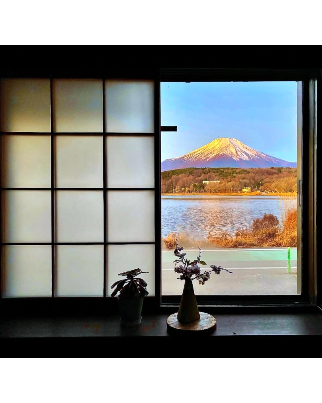 View of Mount Fuji through a window, with traditional Japanese shoji screen on the left, a vase with flowers, and a plant on the windowsill.