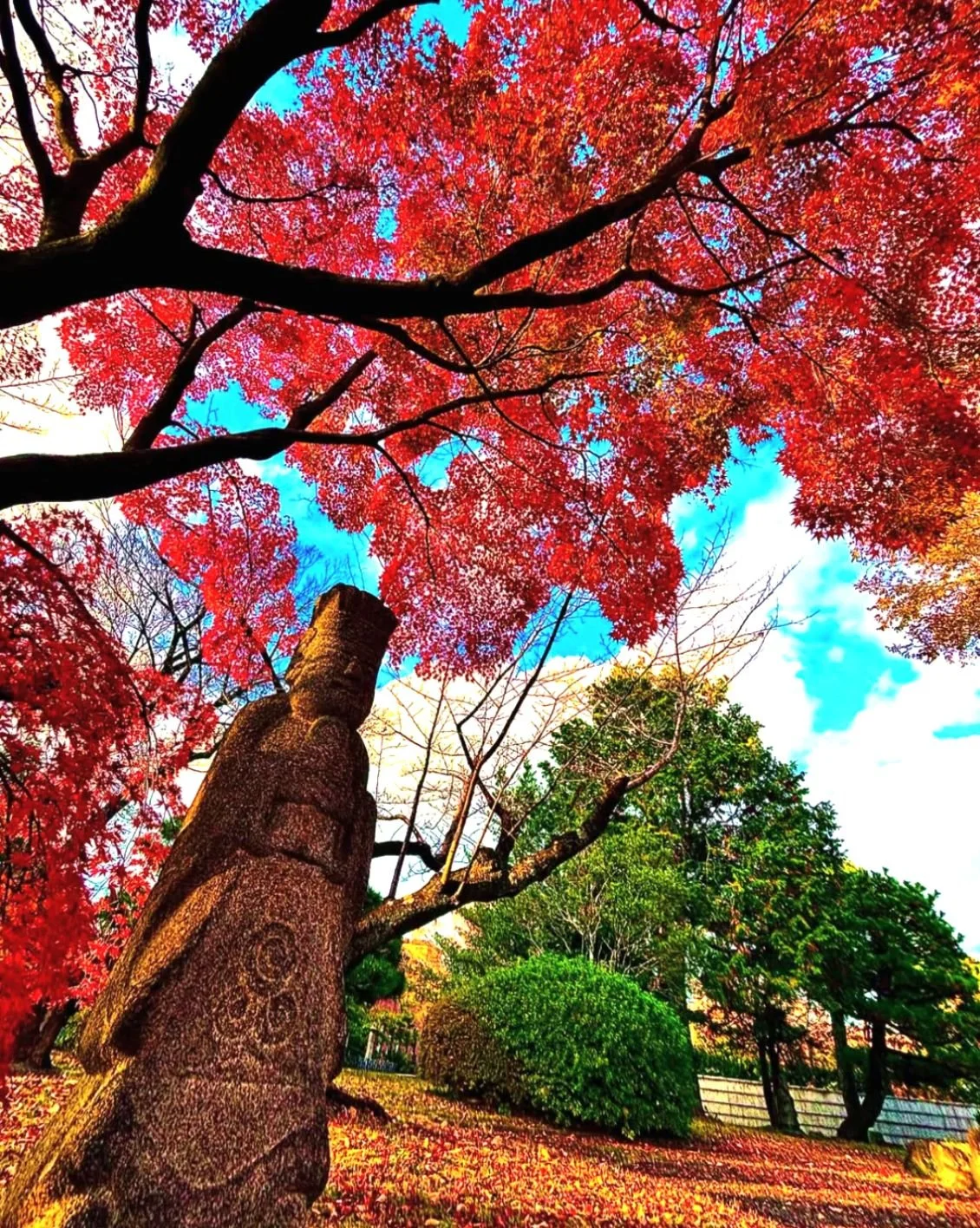 Colorful autumn scene with red and green trees, a carved stone statue, and fallen leaves on the ground, under a blue sky with clouds.