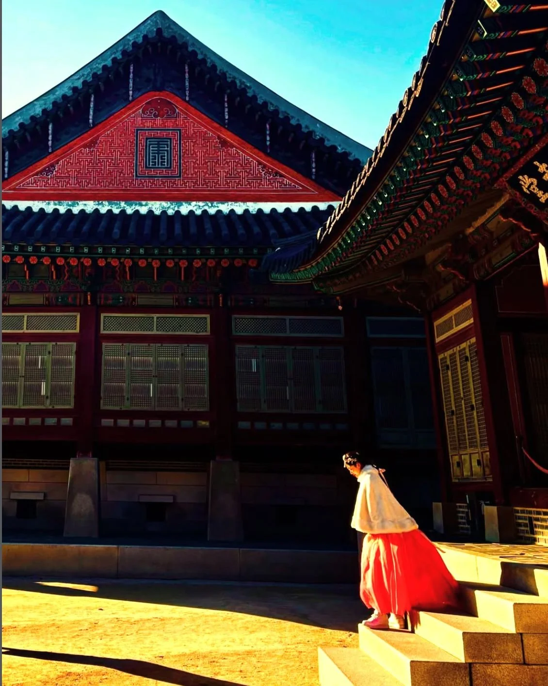 A woman in traditional Korean hanbok walking down stone steps outside a traditional Korean building with ornate, colorful roof and wooden lattice windows, illuminated by warm sunlight.