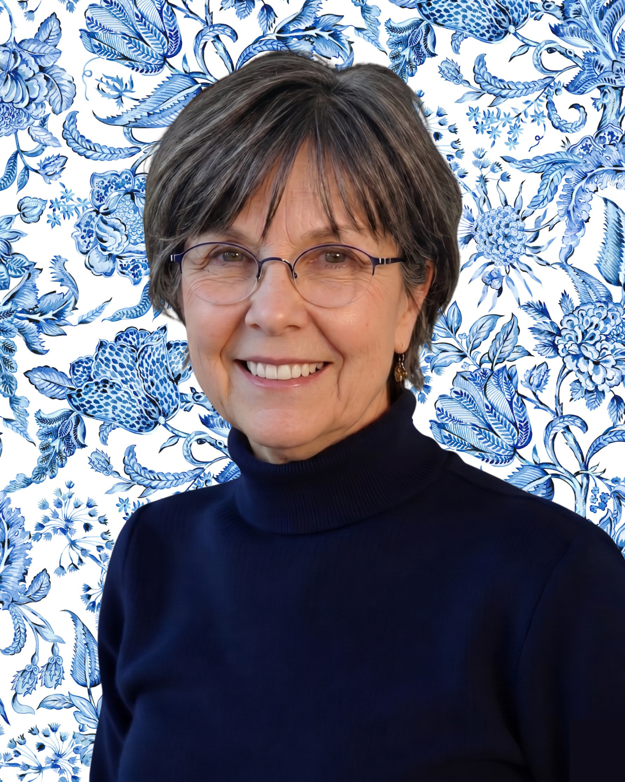 Portrait of a smiling woman with short brown hair, glasses, and wearing a dark turtleneck sweater, standing in front of a blue and white floral patterned background.