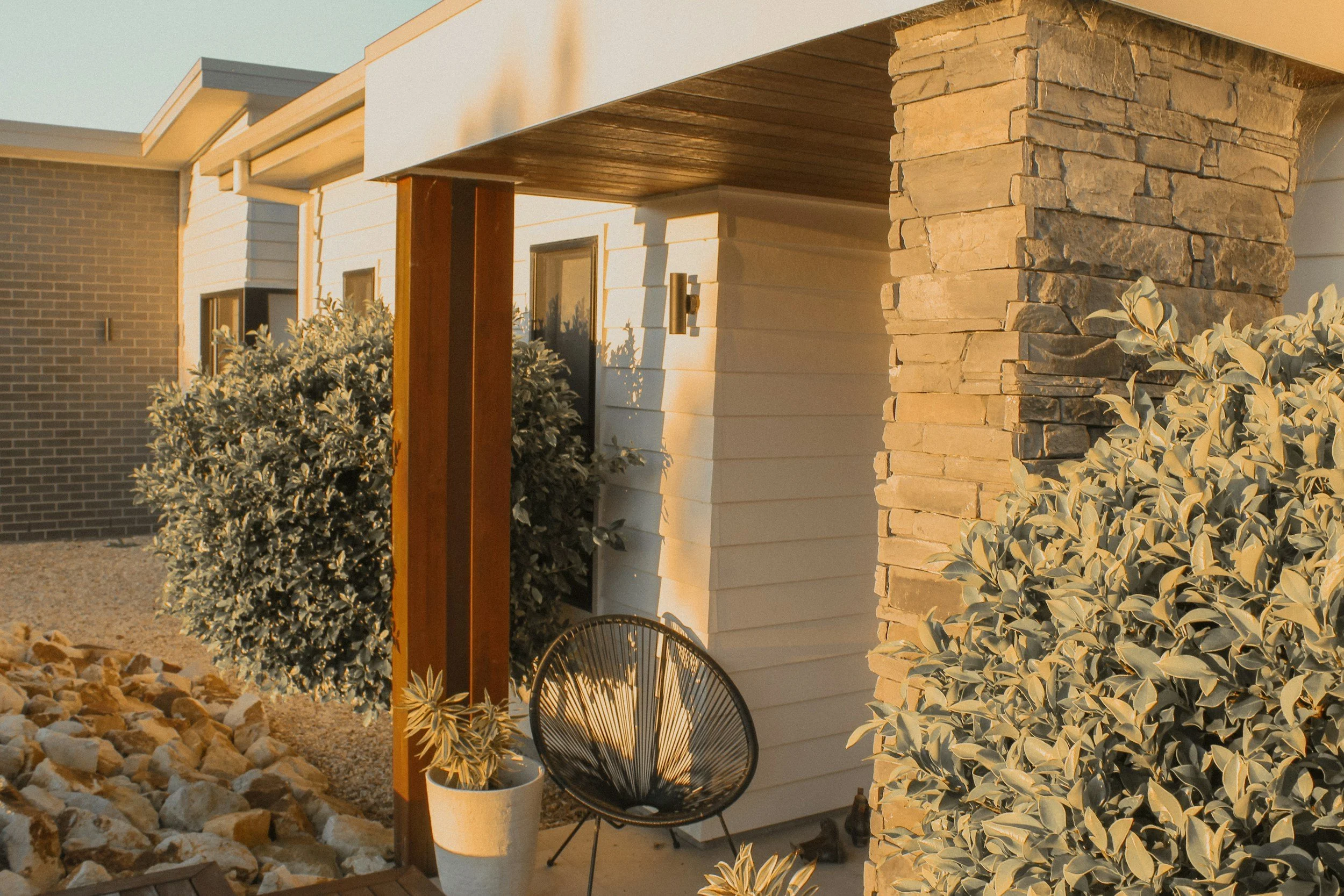 Front porch of a modern house with white siding, a stone column, a wooden support beam, potted plants, a black metal chair, and landscaped rocks and bushes, illuminated by warm sunlight.