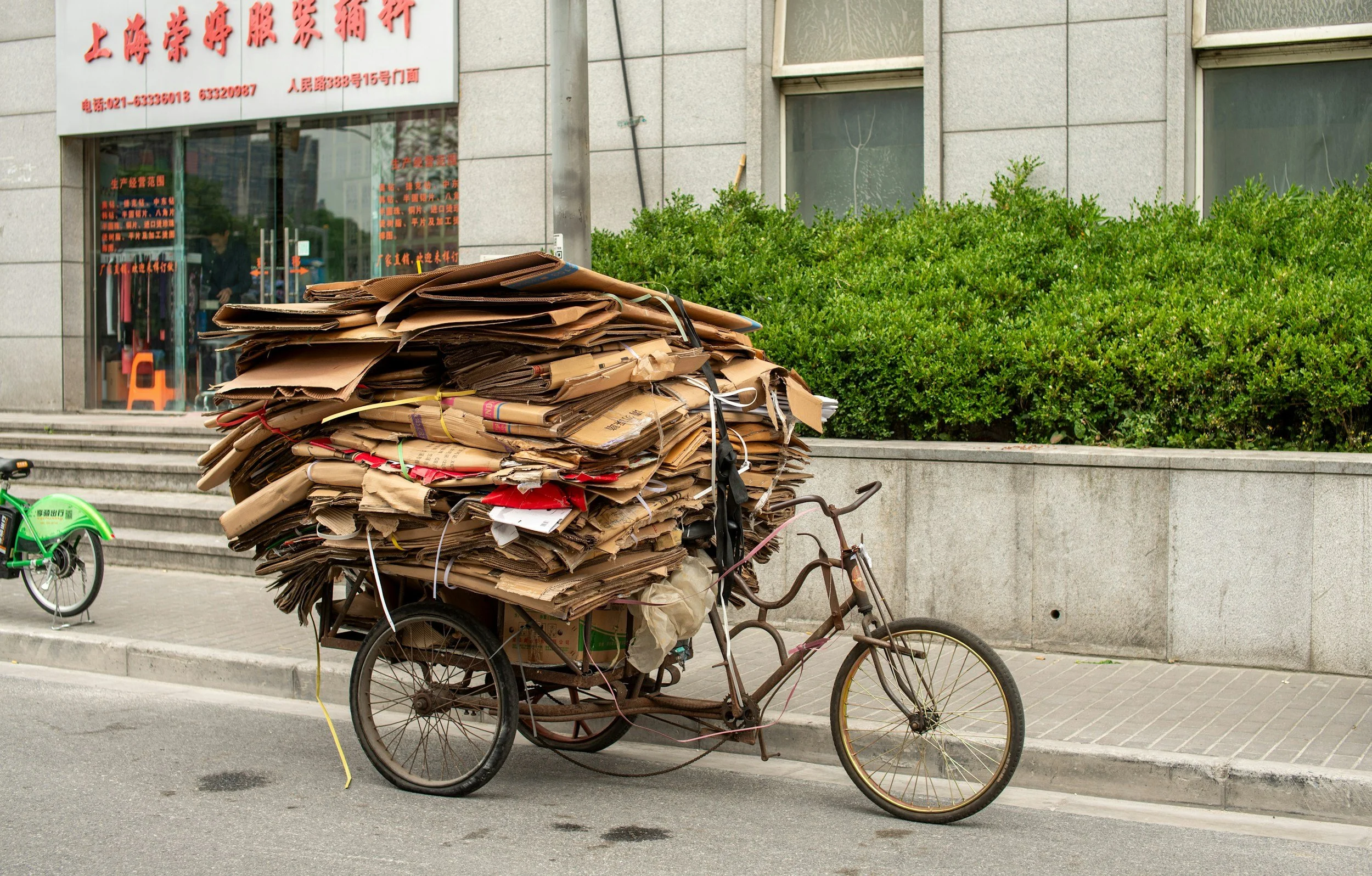 A rusty tricycle cart loaded with a large pile of flattened cardboard boxes parked on a city sidewalk.