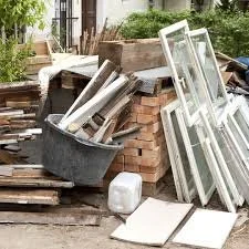 Piles of building materials and tools, including window frames, bricks, a bucket with pipes, and other construction supplies outdoors.