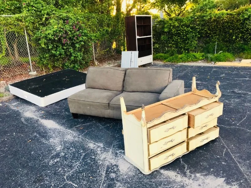 Various furniture items including a gray sofa, a vintage dresser, and a mattress are outdoors on an asphalt surface, with a chain-link fence and green trees in the background.