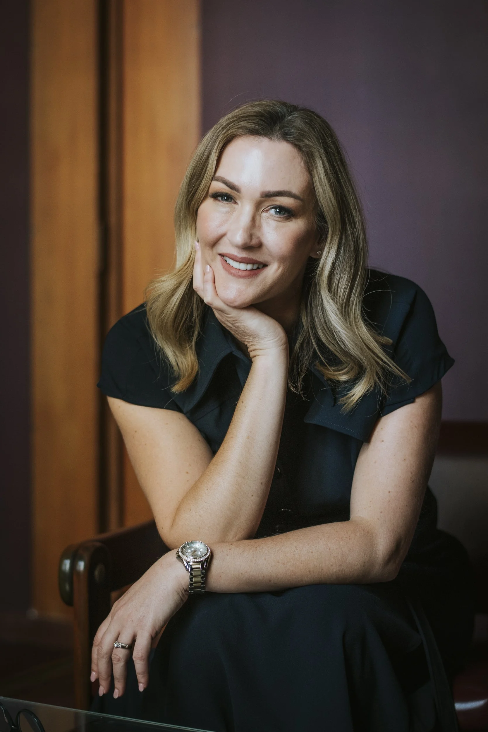 A woman with blonde hair - Barratt Law principal Cara Barratt - smiling, resting her chin on her hand, and wearing a watch and diamond ring, sitting at a table in front of purple and orange curtains.