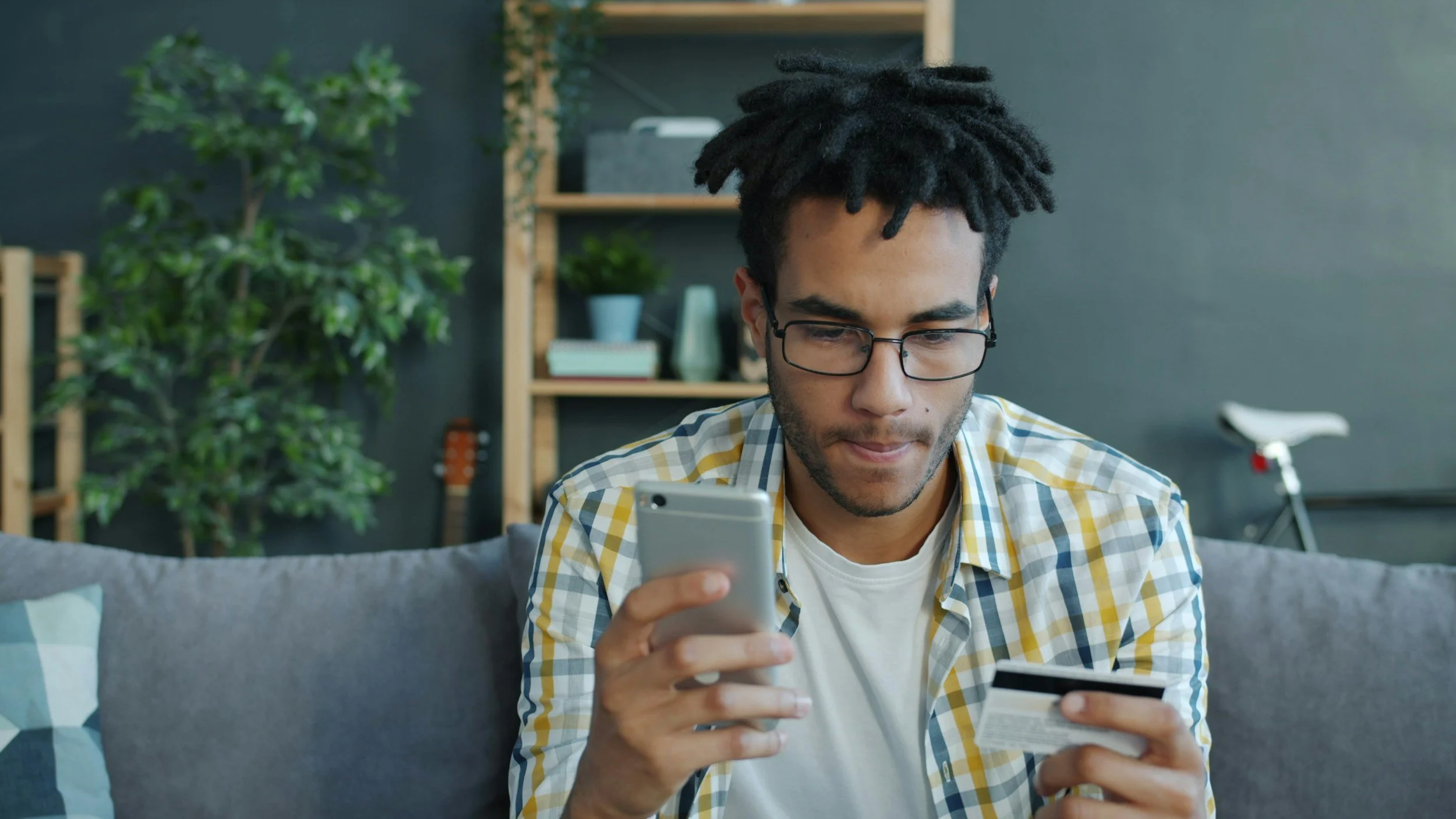 Young man with glasses and a plaid shirt sitting on a grey couch, looking at his smartphone while holding a credit card in his other hand inside a modern living room with plants, books, a guitar, and a bicycle in the background.