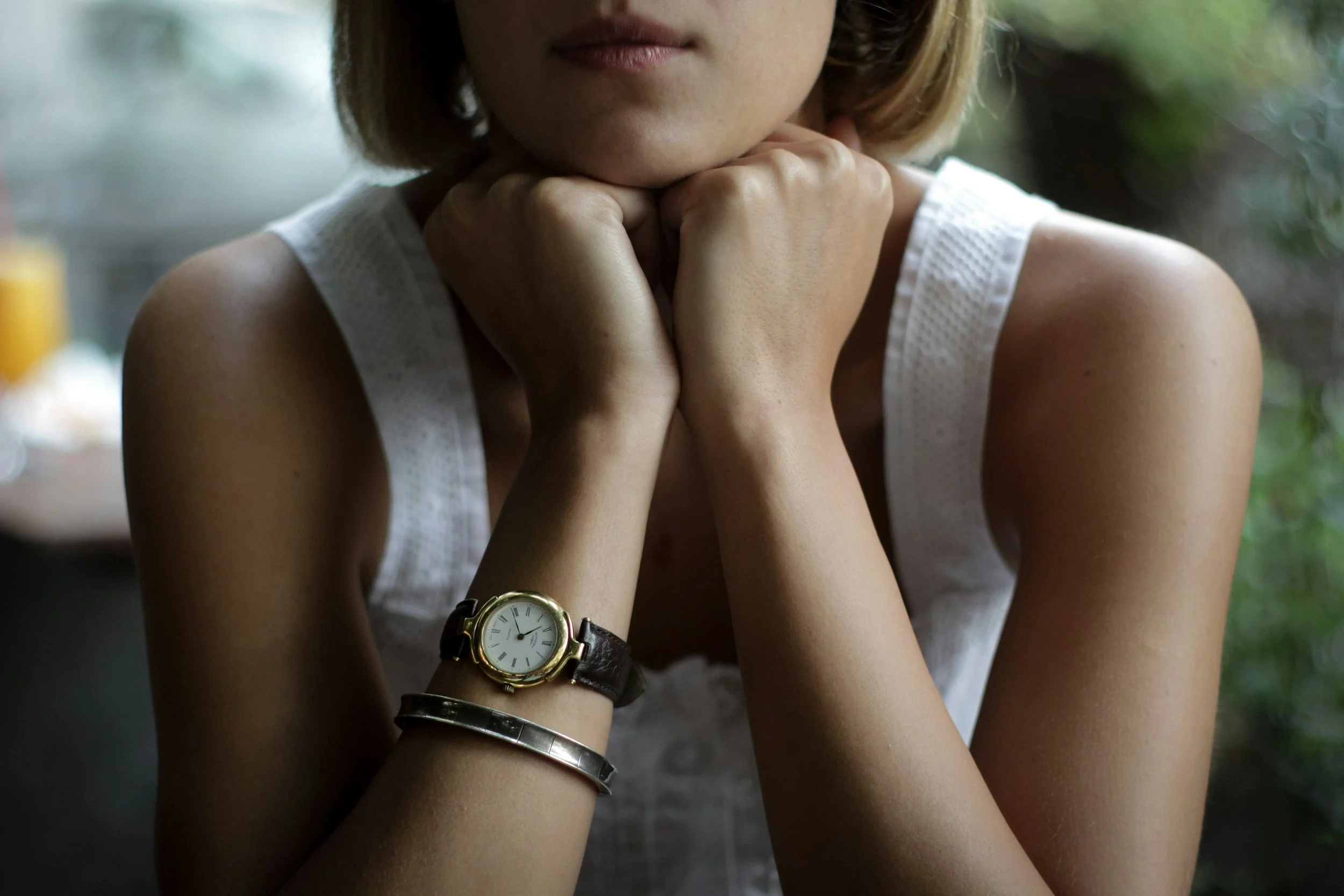 A woman with short hair rests her chin on her hands, which are crossed over her knees. She wears a white sleeveless top, a gold watch, and silver bracelets. The background is blurred with green foliage.