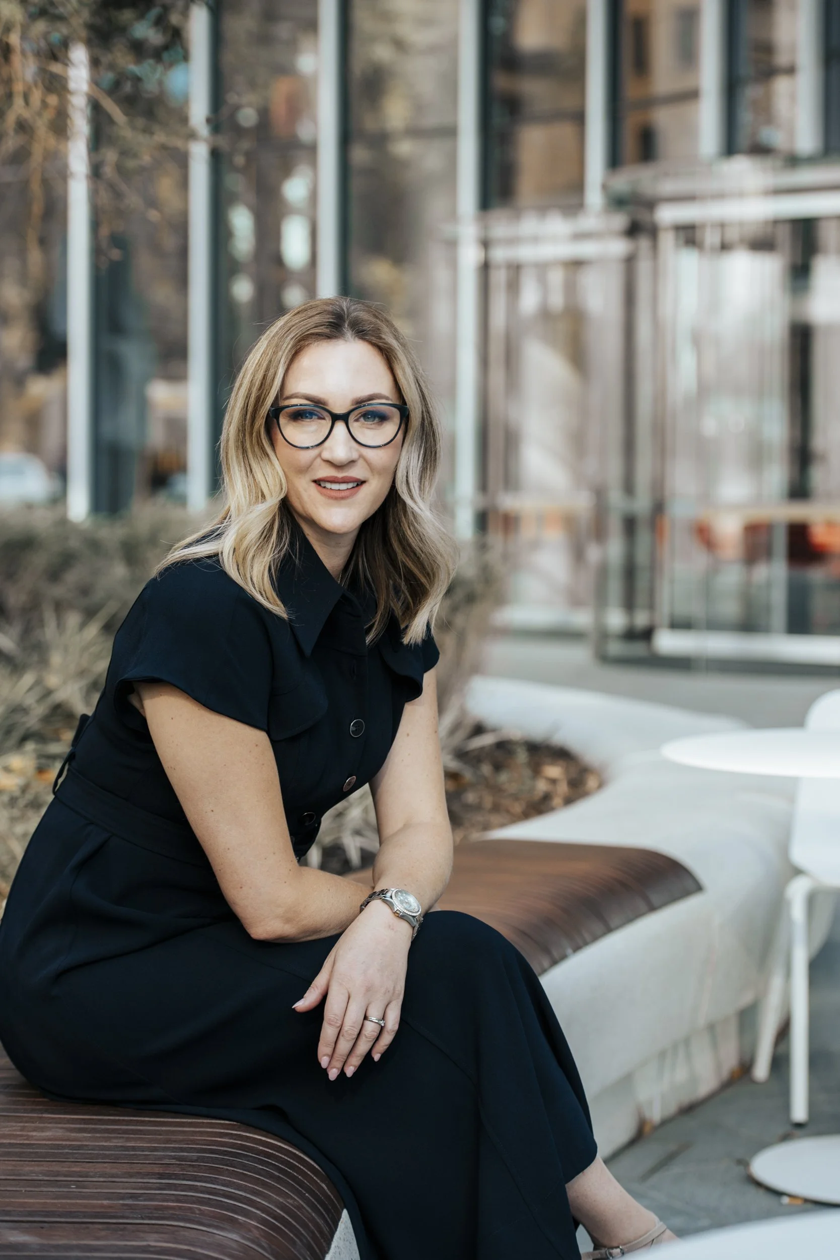 A woman with blonde hair, glasses, and a black dress sitting on a curved wooden and concrete outdoor bench with white tables in the background.