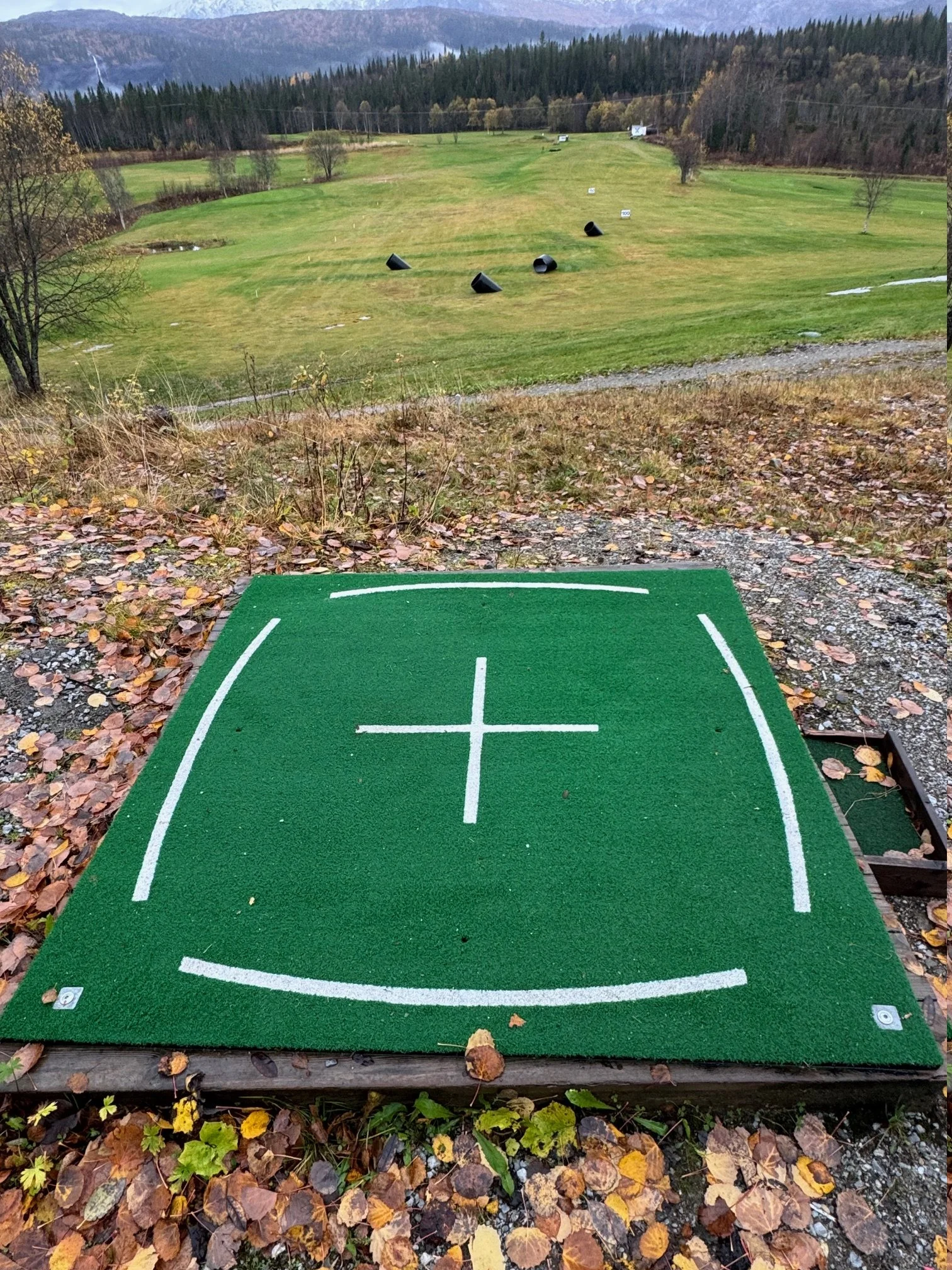 En utendørs minigolfbane med et lite utslagsted, med en grønn matta og en hvit oppmerkning, utsikt over en golfbane med flere baller og kjegler, omgitt av høstløv og trær.