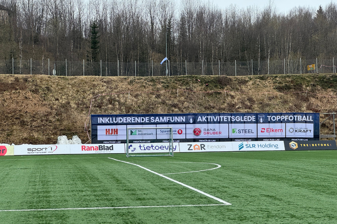 Fotballbane med reklameskilt og gressbakke, omgitt av tre og en skog i bakgrunnen.