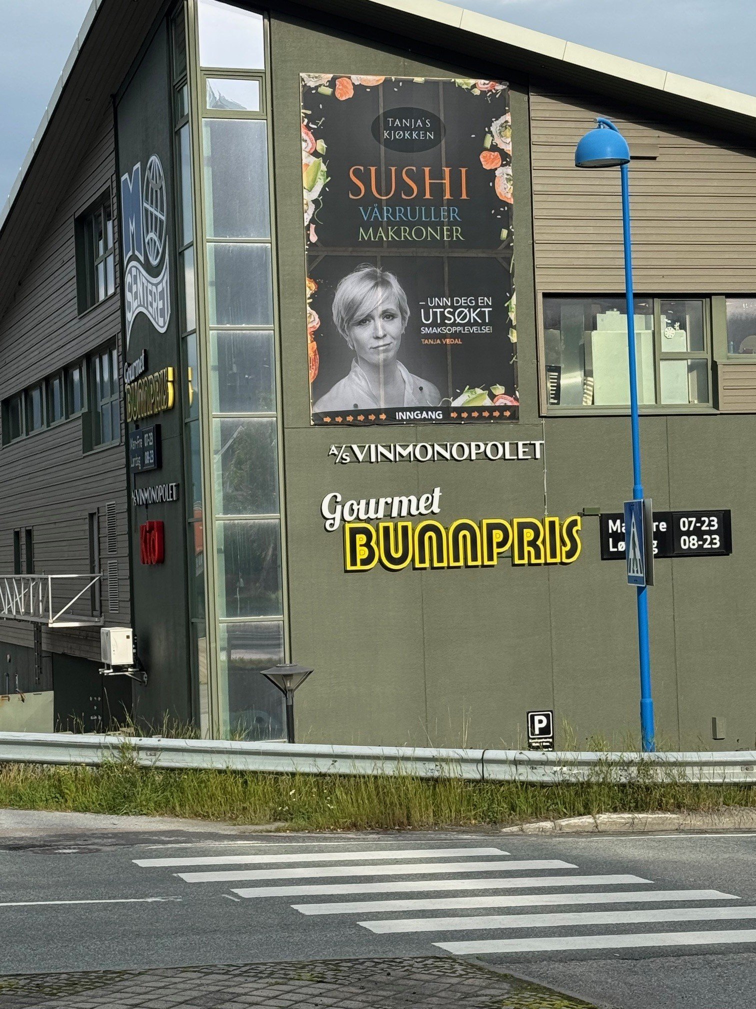 Fasad av kjøpesenter med store reklameskilt for sushi og mat, samt et skilt med åpningstider og parkering