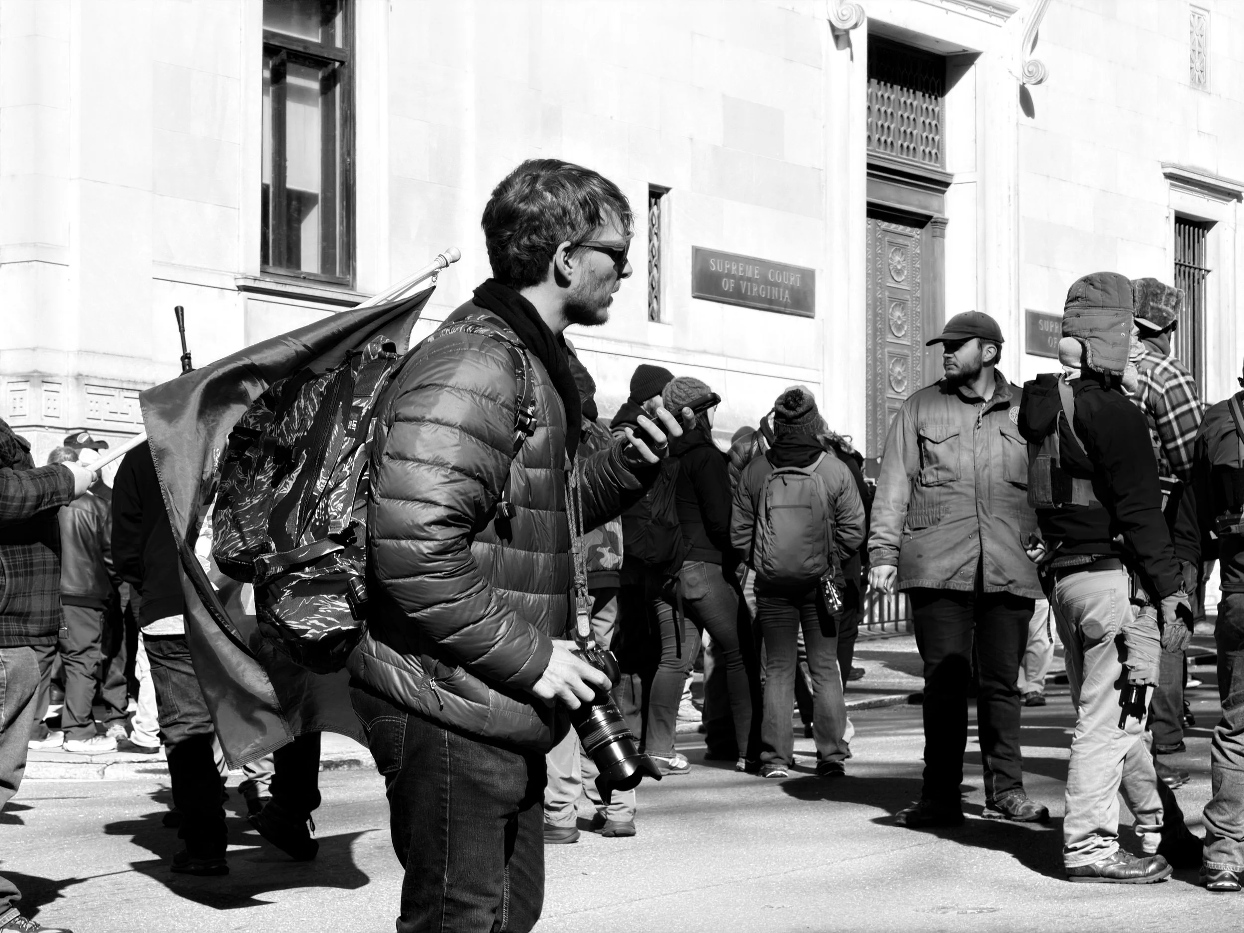 A man with glasses, a camera, and a backpack holding a phone, standing among a crowd outside a government building.