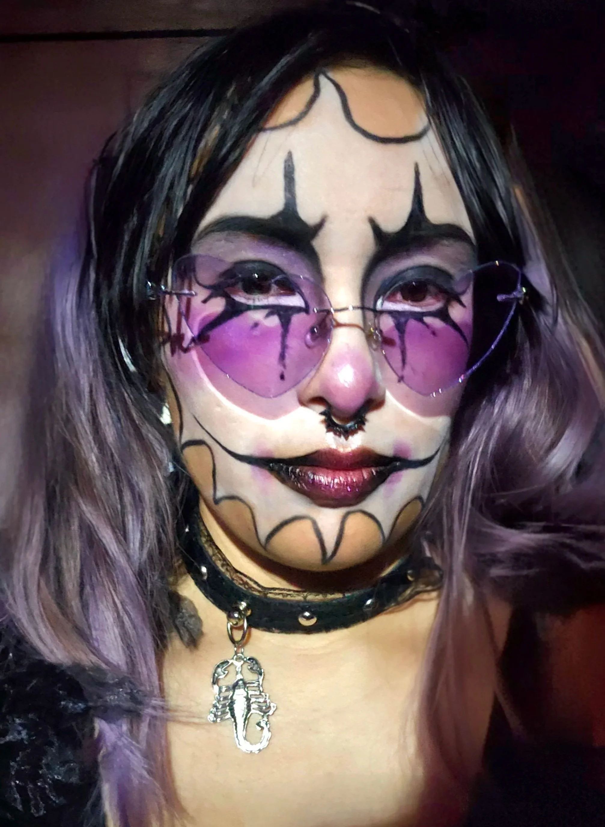 A woman with purple hair and gothic makeup, wearing pink heart-shaped sunglasses with black bat wing designs, a black choker, and a necklace with a skeleton key charm.