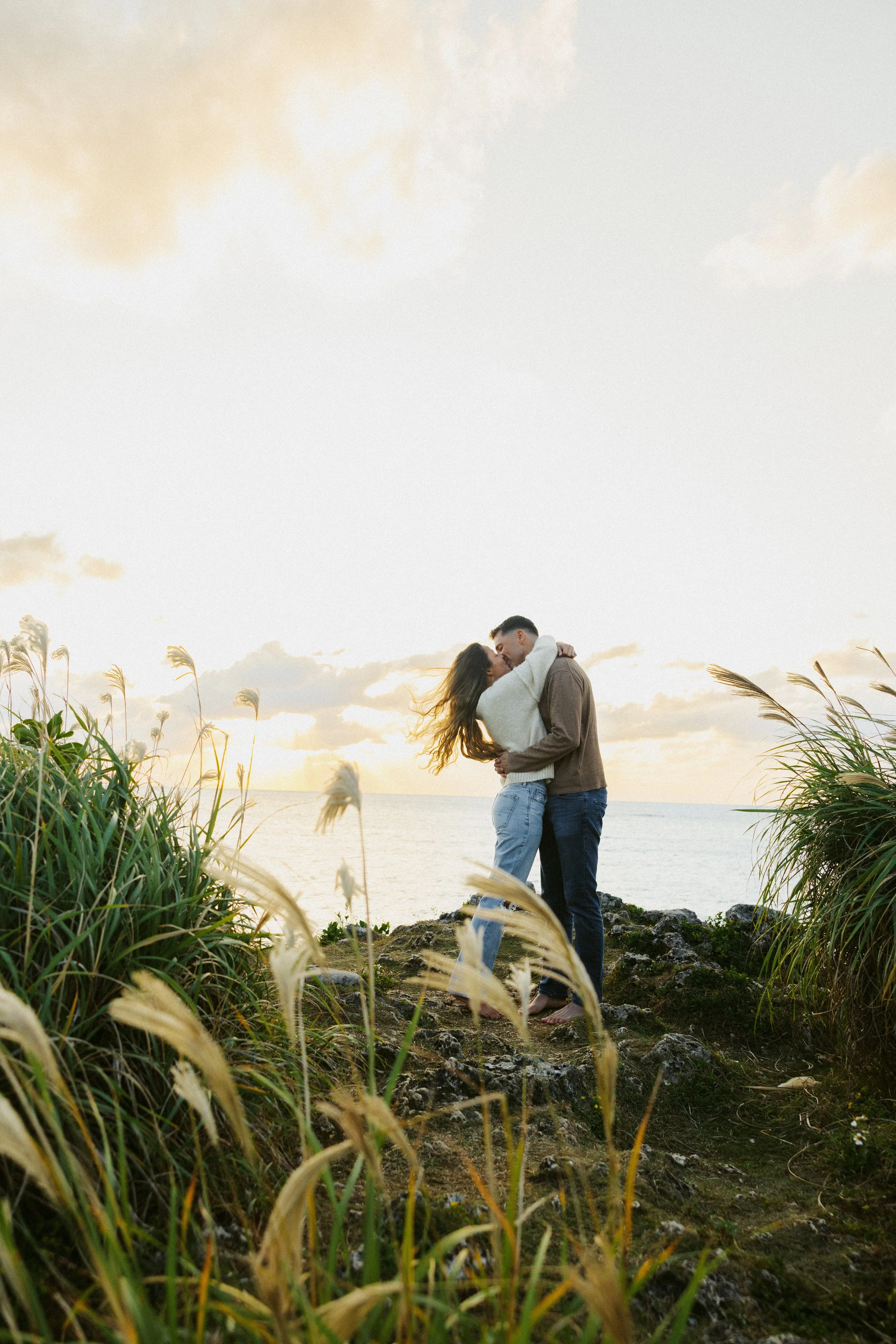 A couple embracing and kissing near the ocean at sunset, surrounded by tall grass and rocky terrain.