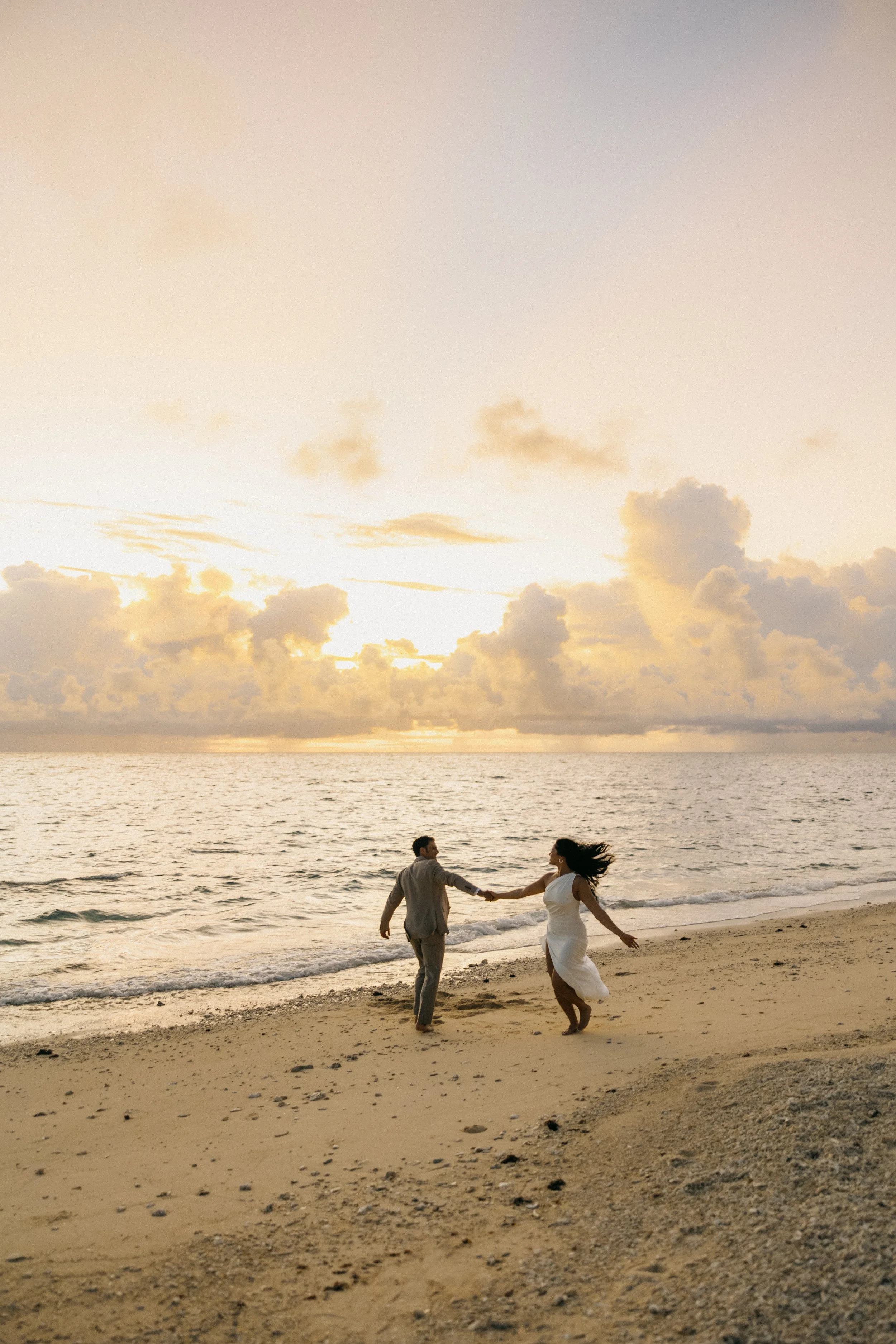 A couple dancing on the beach during sunset, holding hands, with the ocean and cloudy sky in the background.