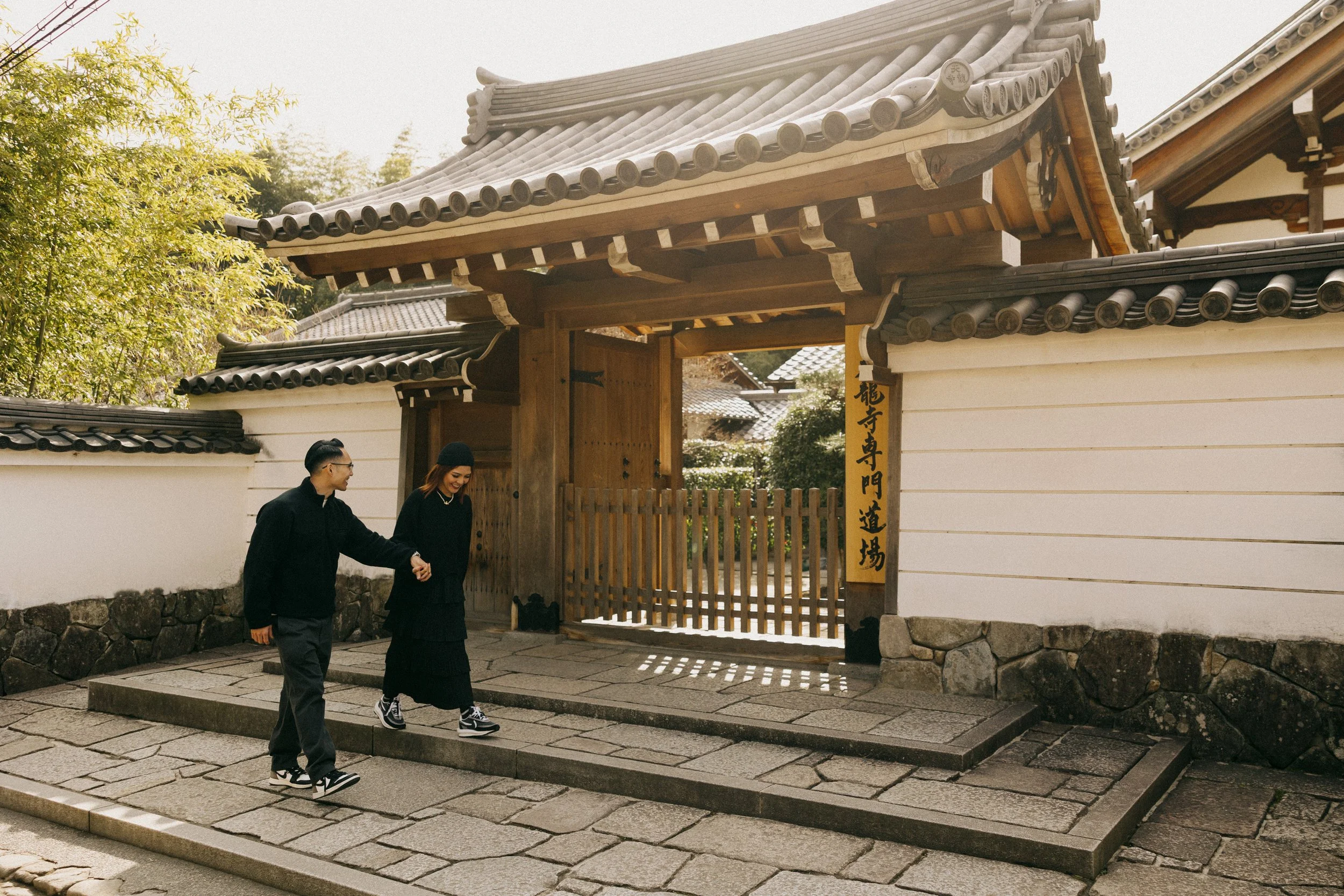 Two people walking hand in hand through a traditional Japanese temple gate with tiled roof and wooden accents, surrounded by stone pavement and natural greenery.