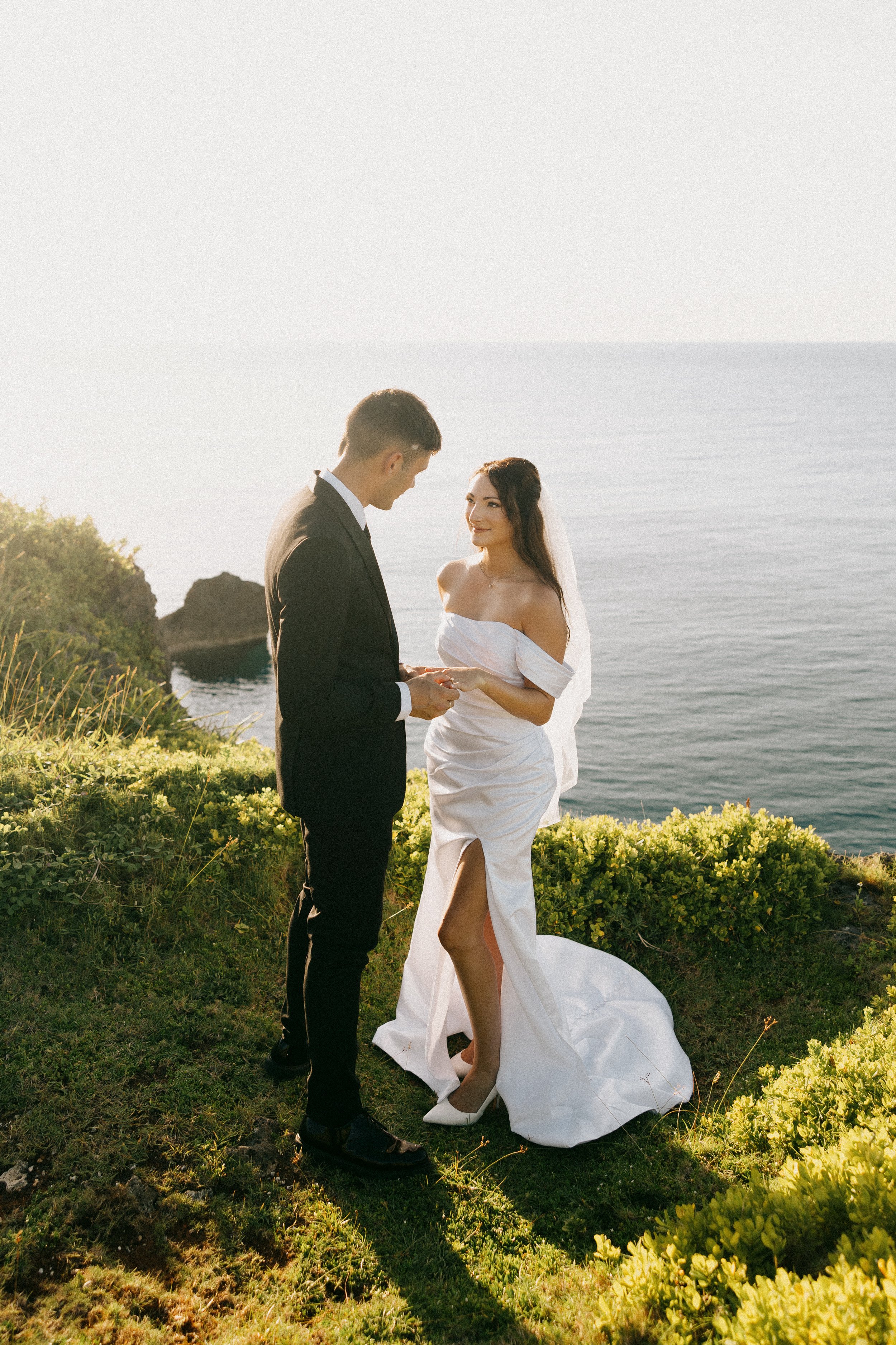 A couple in wedding attire exchanging vows outdoors by the ocean at sunset, with greenery and rocky cliffs in the background.