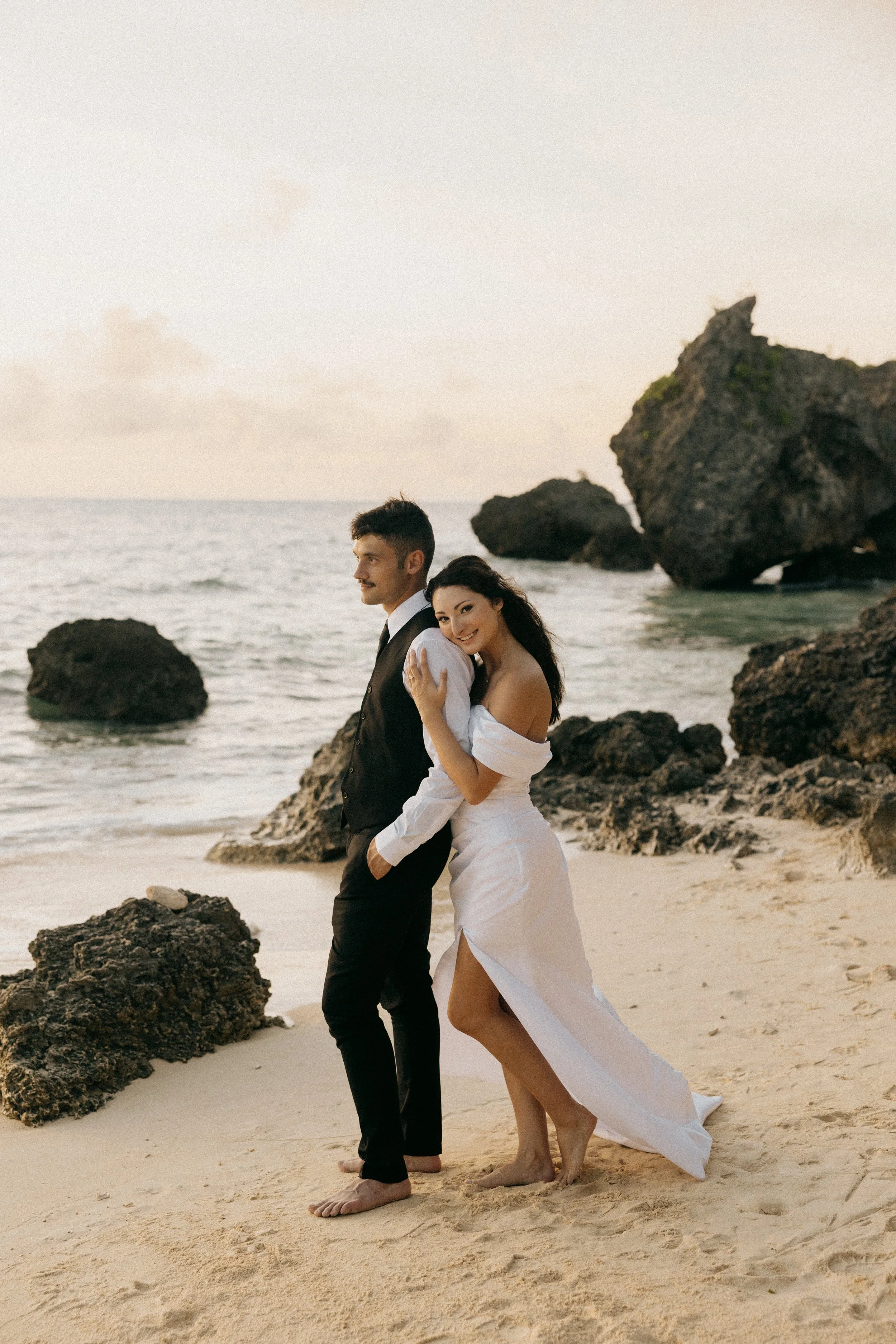 A couple wearing wedding attire standing barefoot on a sandy beach with large rocks and the ocean in the background during sunset.