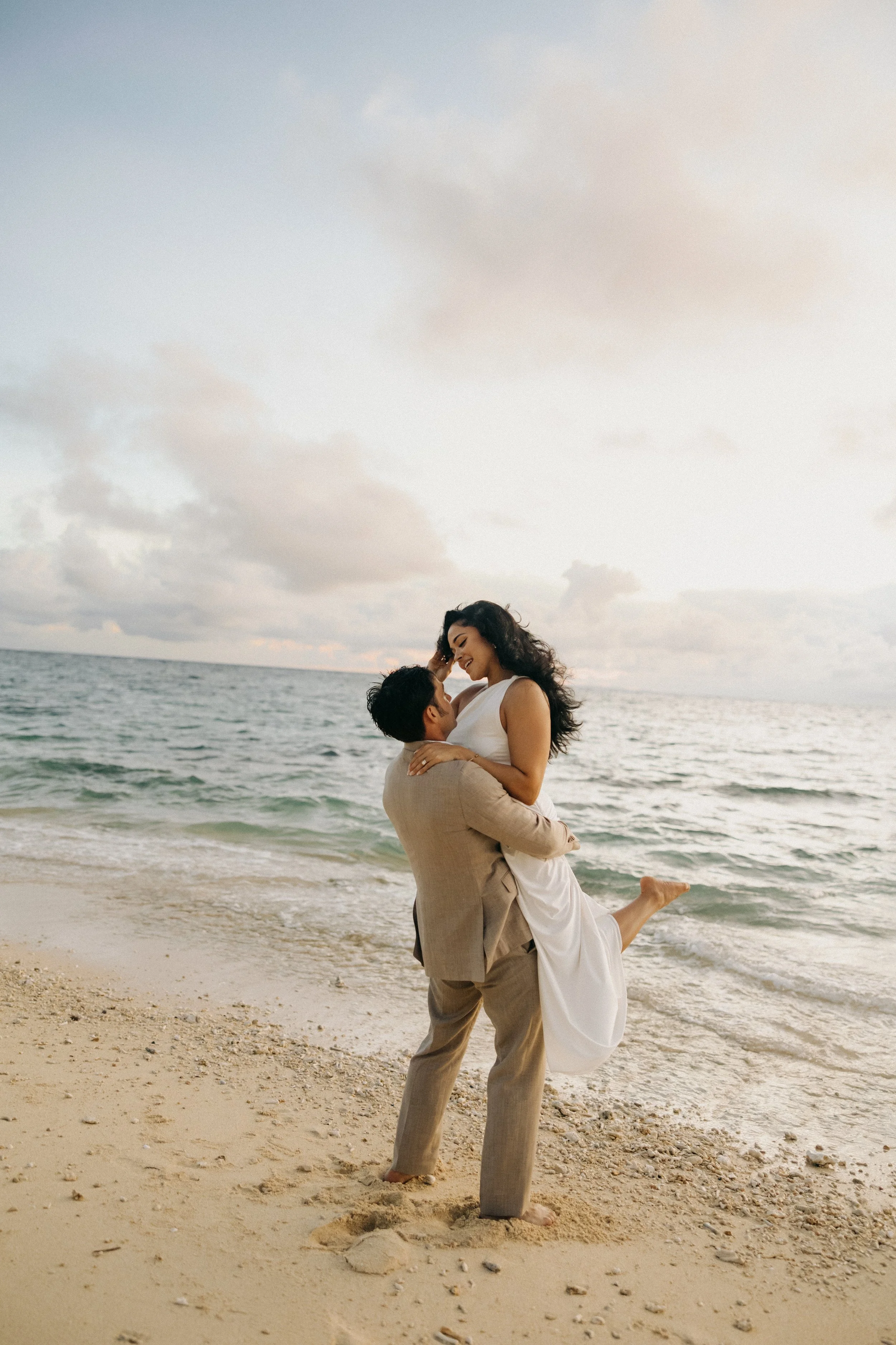 A man in a beige suit lifts a woman in a white dress on a sandy beach near the ocean, both smiling and looking at each other as the sun sets or rises, with clouds in the sky.