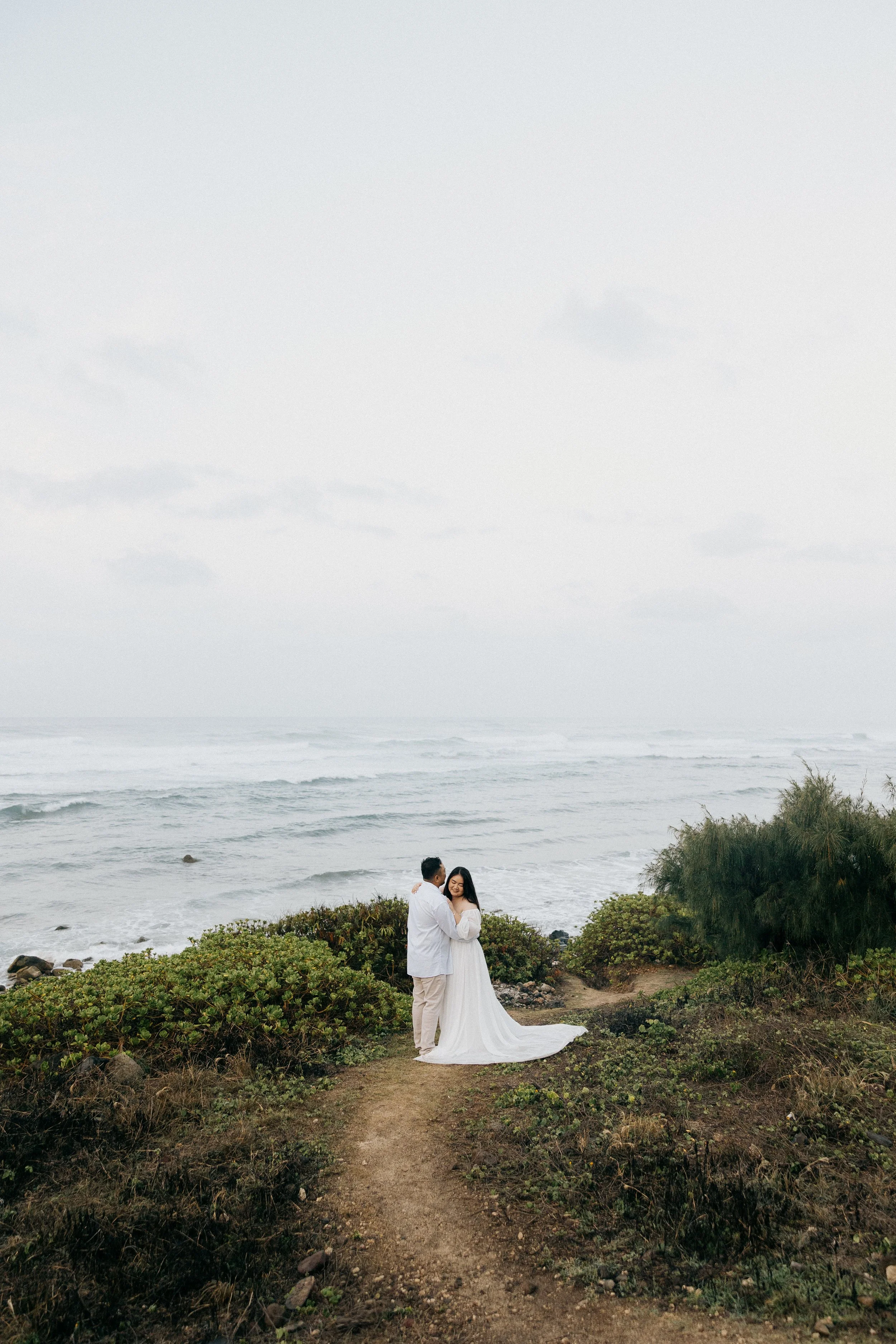Couple dressed in wedding attire standing on a path near the ocean, surrounded by bushes and greenery.