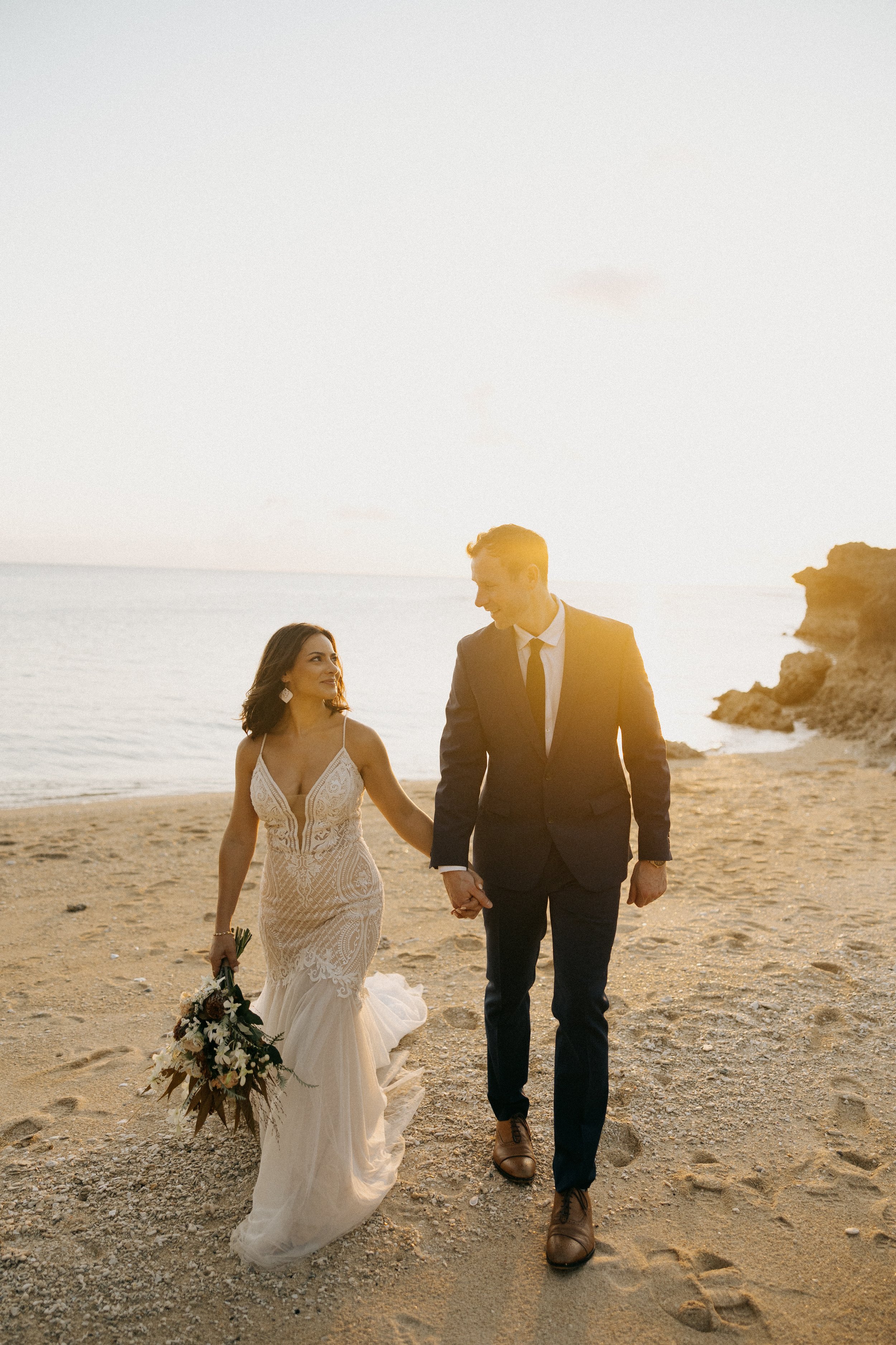 A bride and groom walking hand-in-hand on a beach during sunset, with the bride holding a bouquet of flowers.
