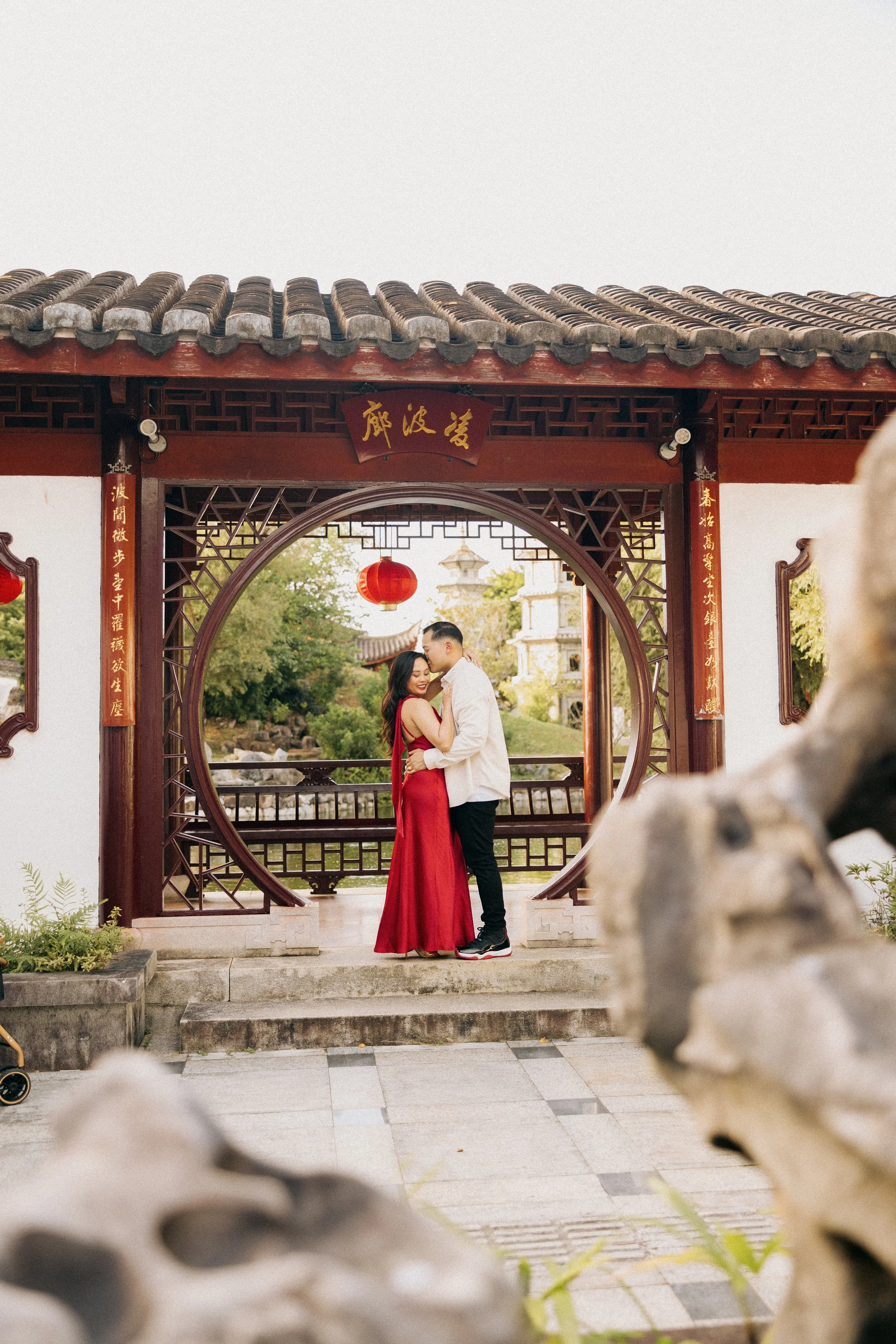 A couple dressed in formal attire, with the woman in a red dress and the man in a white shirt and black pants, standing close together and embracing in front of a traditional Chinese architectural gate with Chinese characters and red lanterns. They a