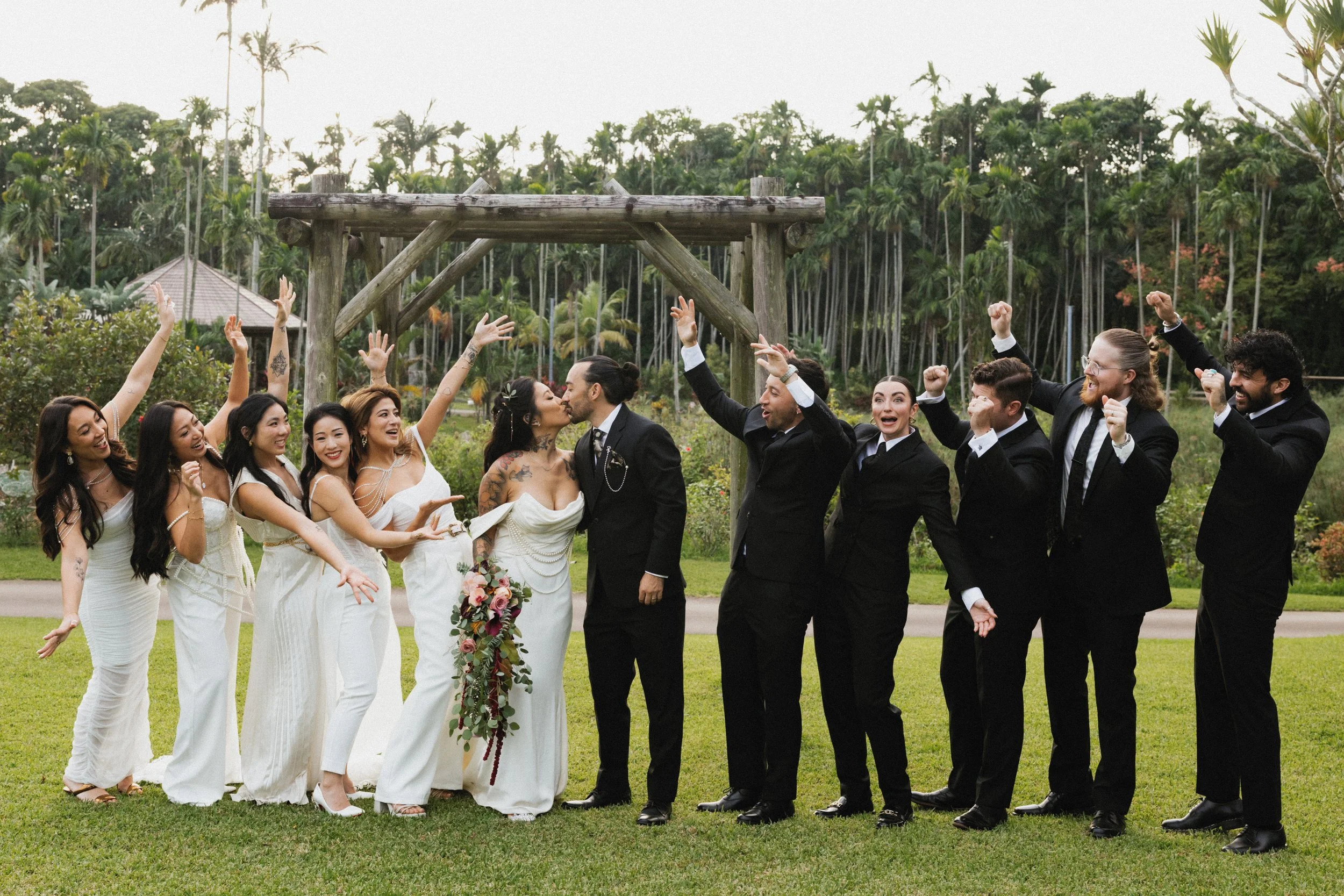 A wedding party of nine people, six women and three men, celebrating outdoors in front of a wooden arch with a lush green background. The bride and groom are kissing, with the bride holding a floral bouquet, and the guests are cheering with their arm