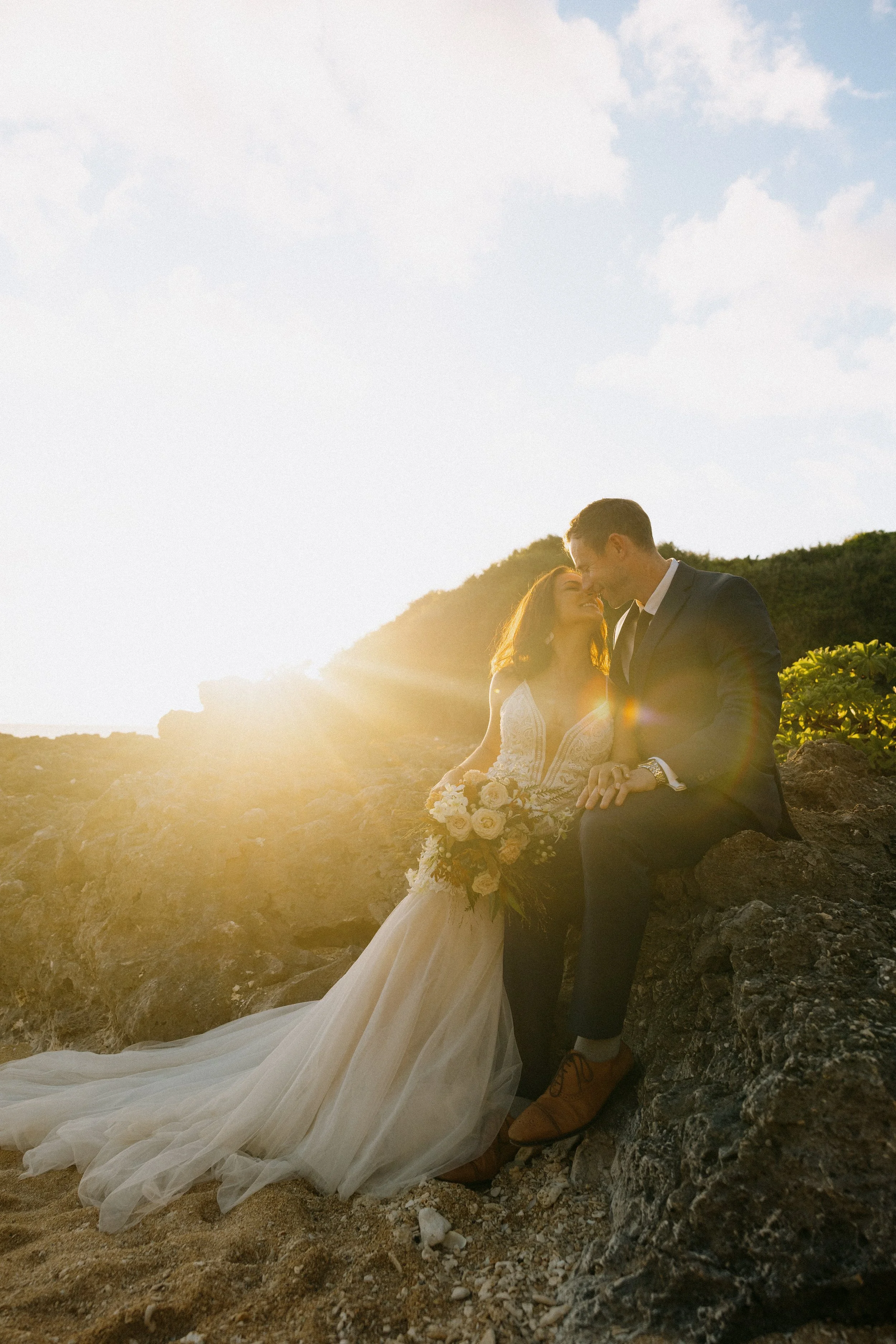 A bride and groom sitting on rocks outdoors at sunset, sharing a kiss, with the bride holding a bouquet of white roses and wearing a wedding dress, the groom in a suit, on a rocky landscape with hills and a partly cloudy sky.