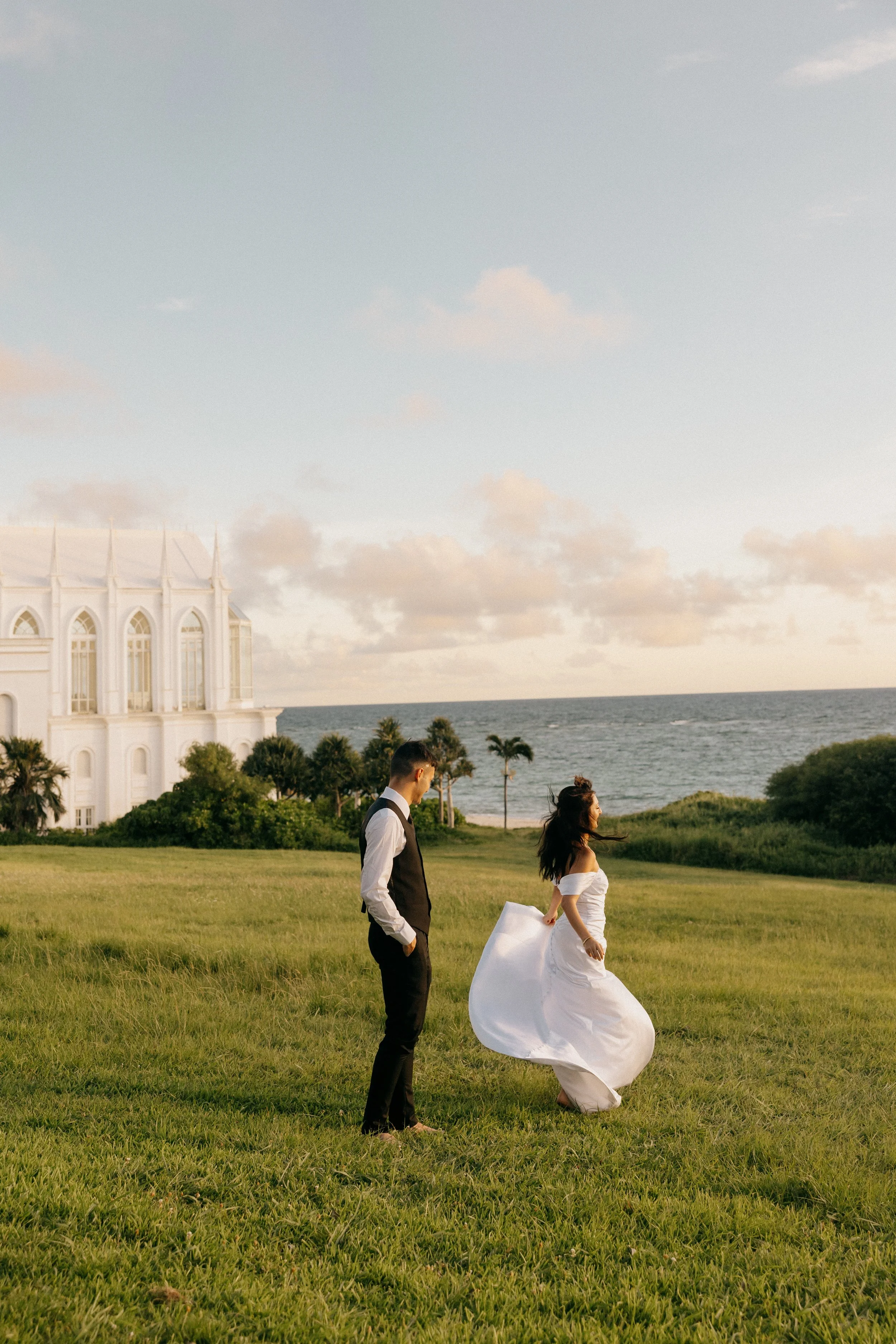 A bride in a white wedding dress and a groom in a black vest and white shirt standing on a grassy field near the ocean, with a white building and palm trees in the background during sunset.