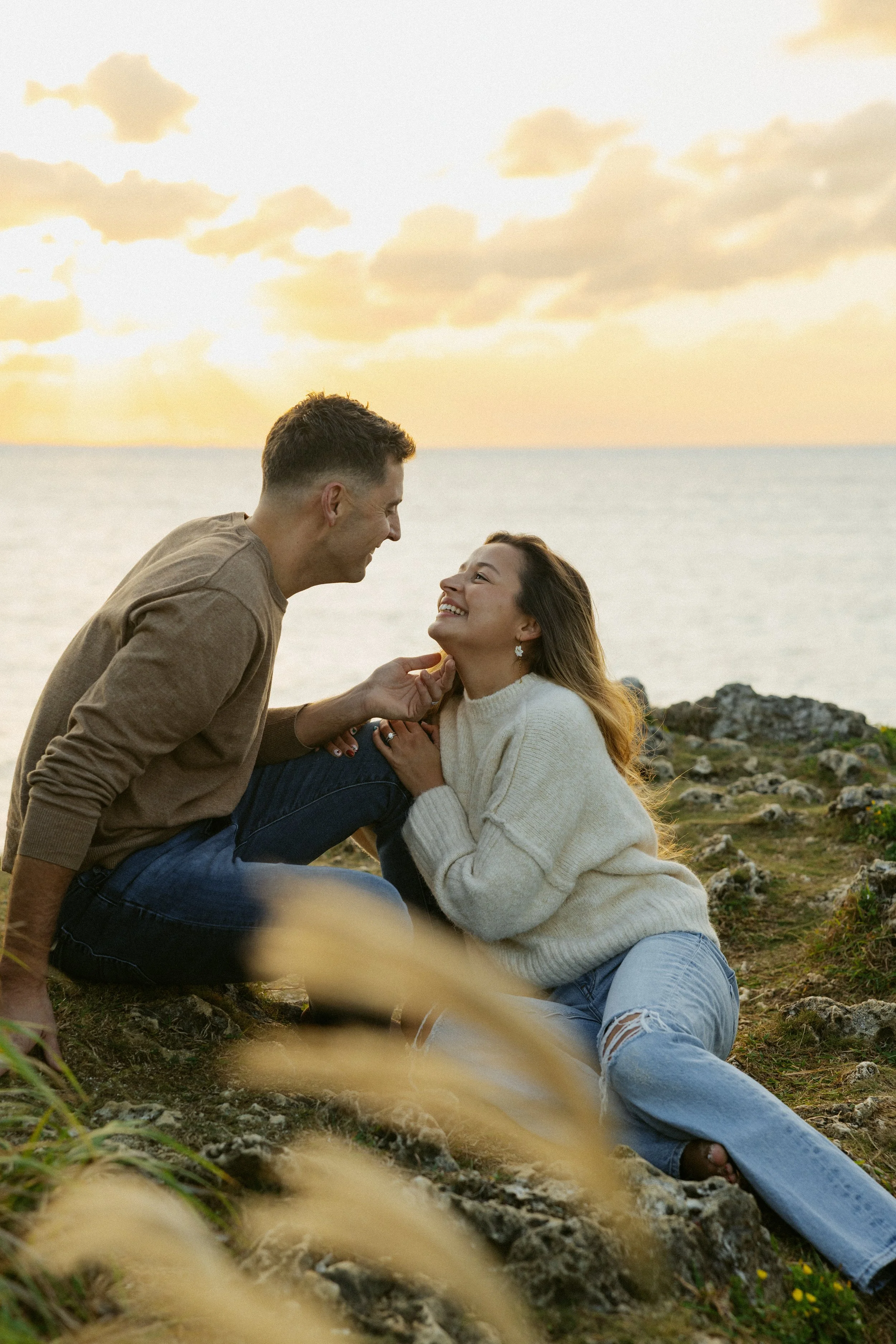 A couple exchanging smiles on a rocky beach at sunset, with the sea and sky in the background.