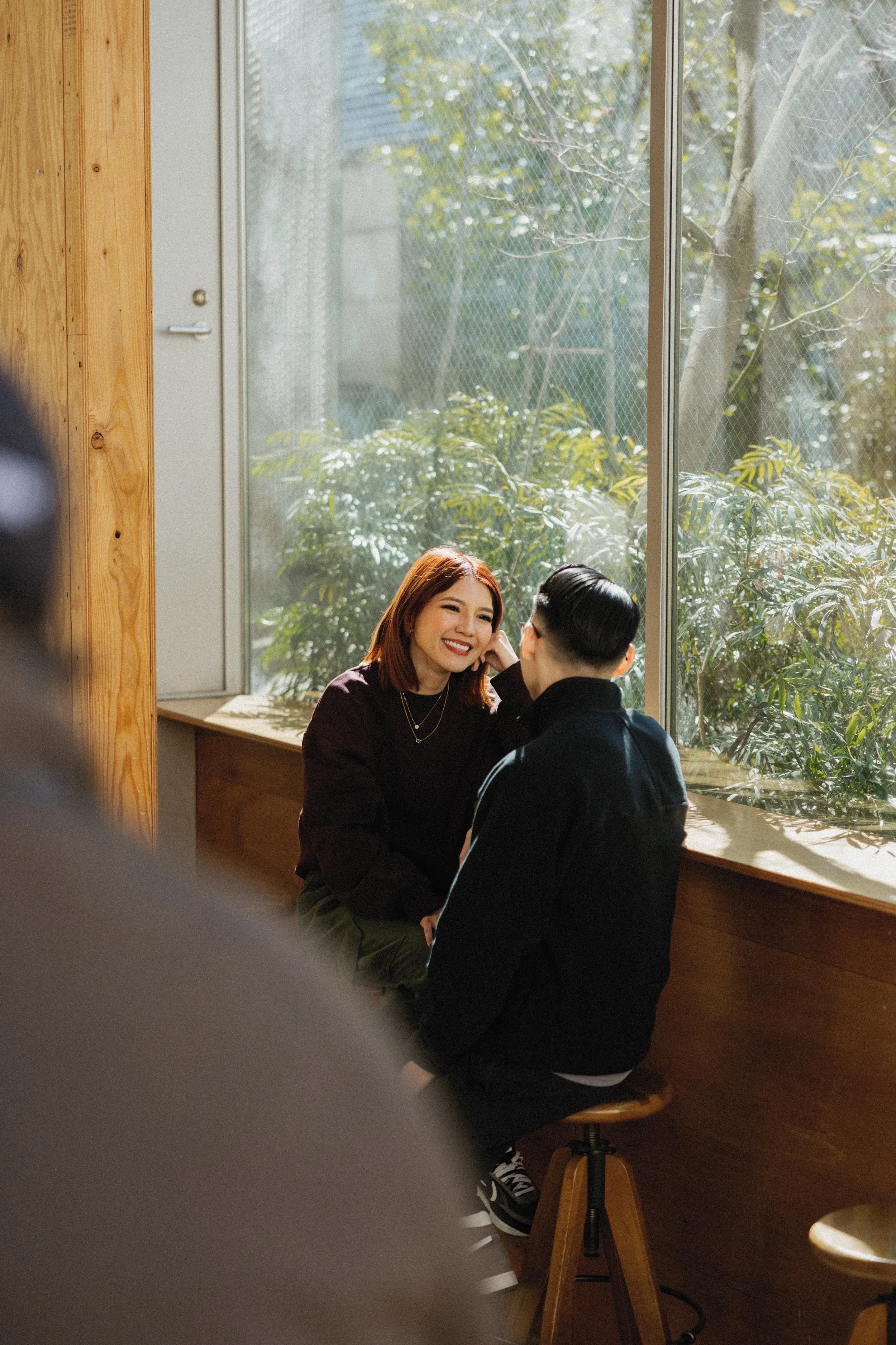 Two women sitting by a large window, smiling and talking, with greenery outside.