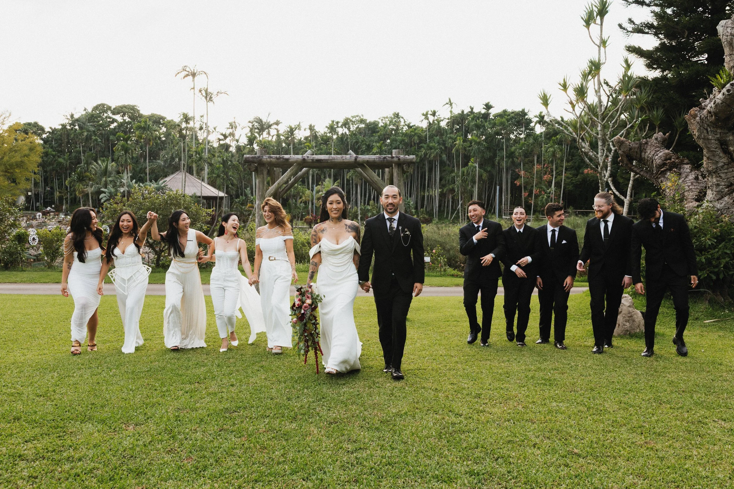 A diverse wedding party walking on a grassy field, with the bride in a white wedding dress and the groom in a black suit at the center, holding hands. Bridesmaids in white dresses are on the left, and groomsmen in black suits are on the right, all sm