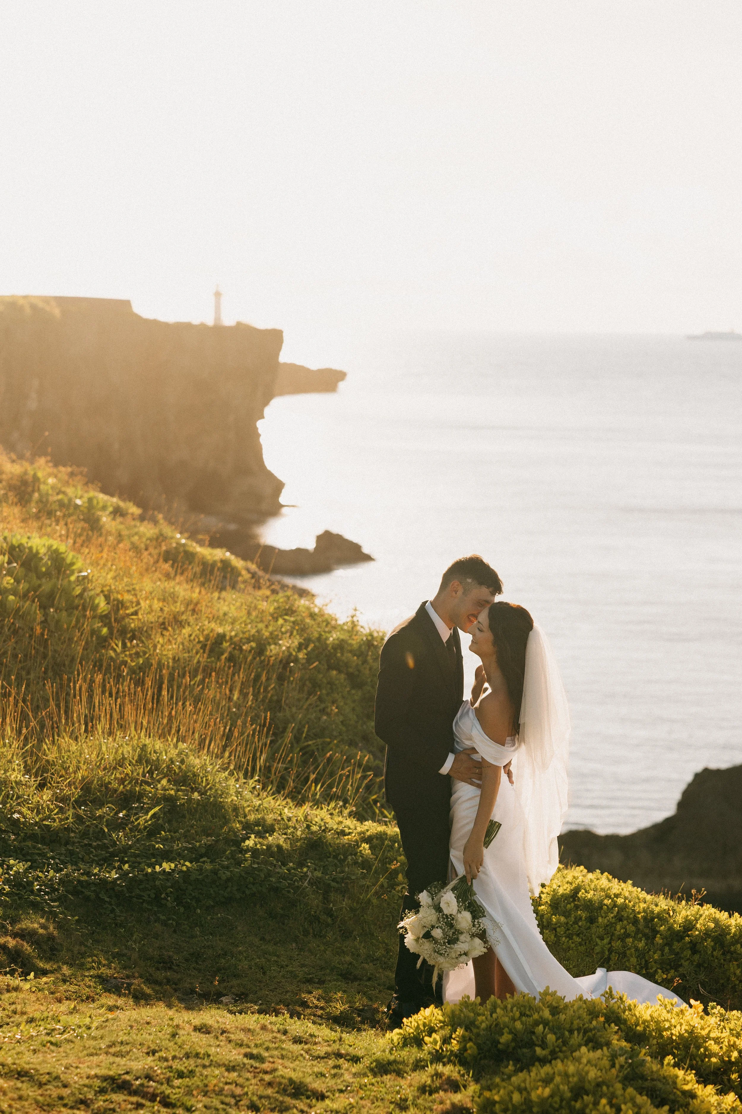A newlywed couple dressed in wedding attire embracing outdoors during sunset near a cliff and ocean.