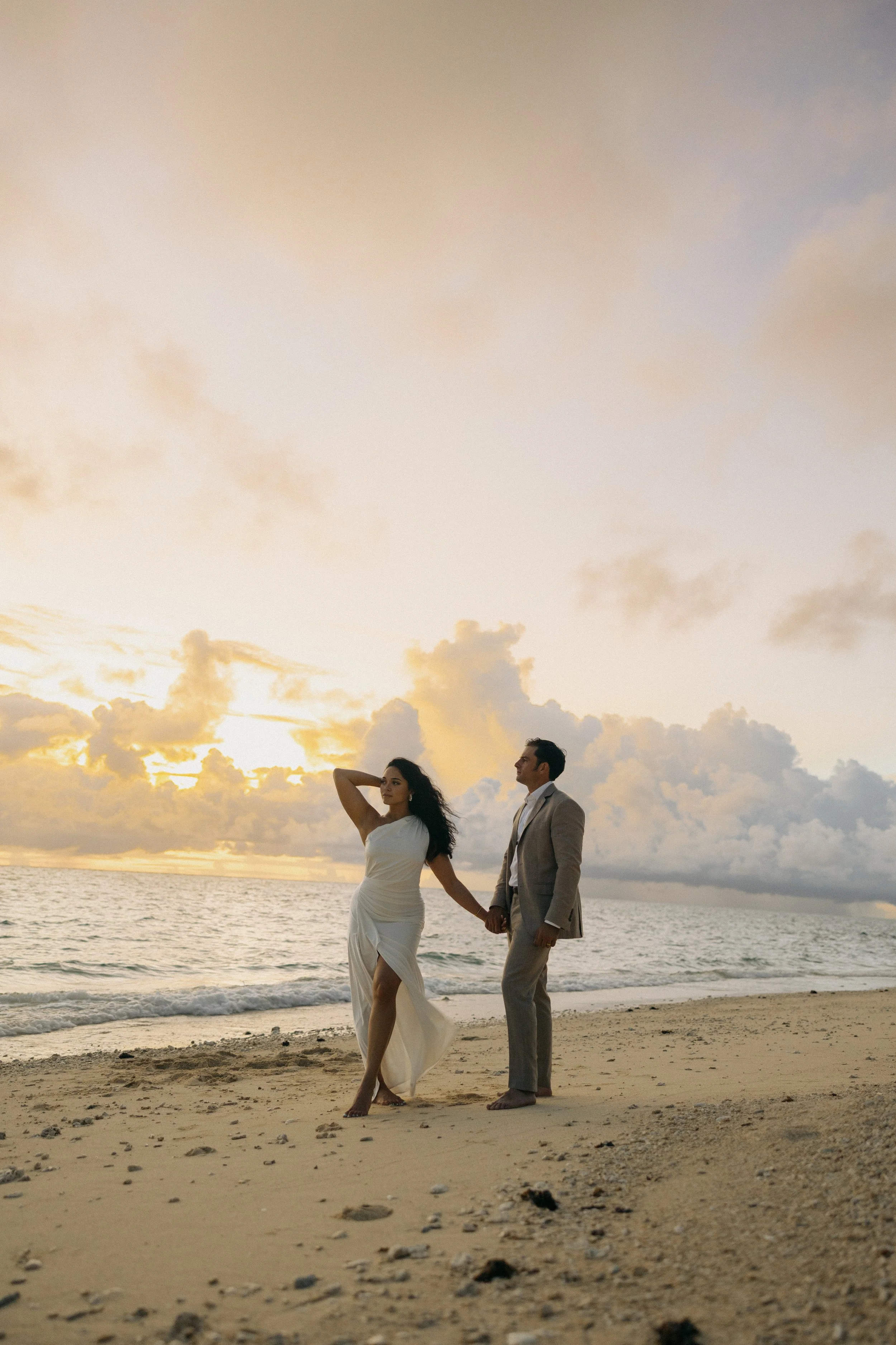 A couple dressed in formal attire, holding hands, walking along a beach at sunset with clouds and ocean in the background.