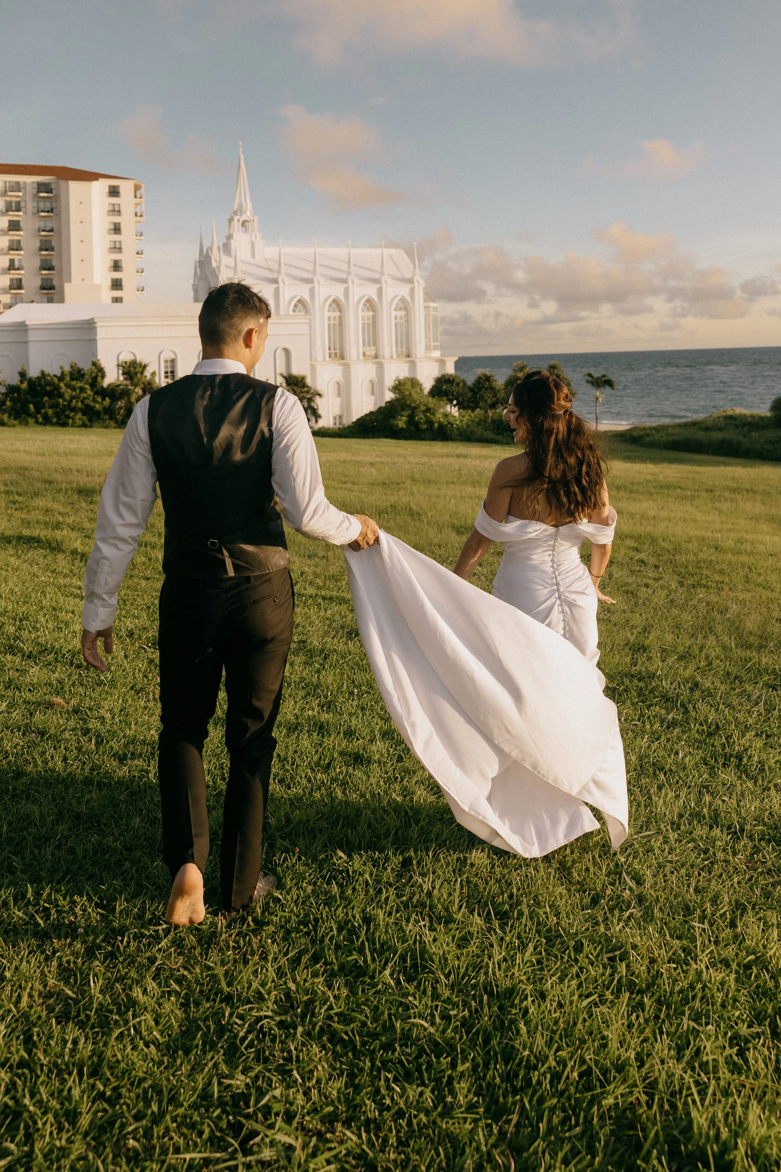 A bride and groom walking barefoot on a grassy field, holding the bride's train, with a white church and ocean in the background at sunset.