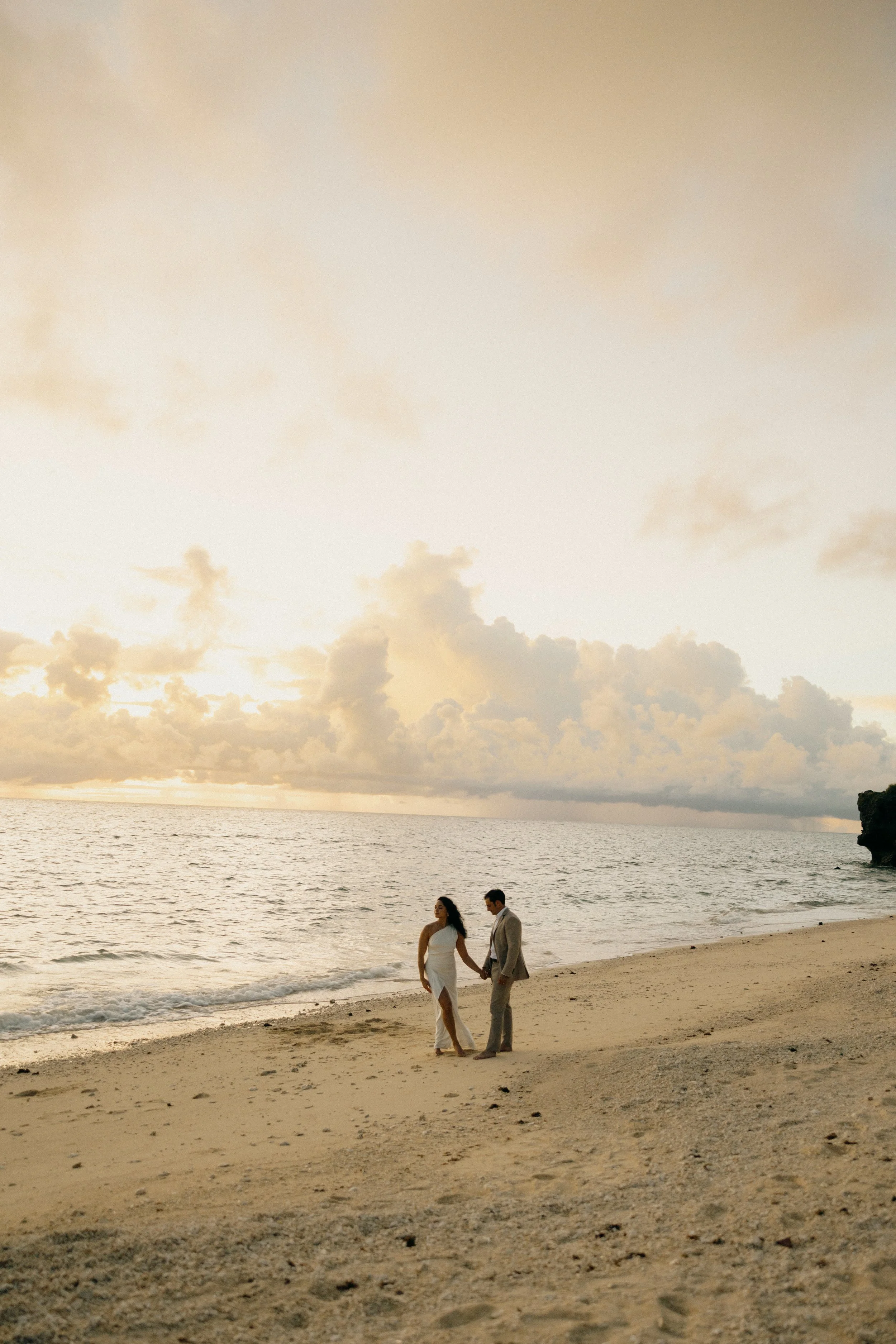 A couple in wedding attire holding hands on a sandy beach during sunset, with clouds and the ocean in the background.