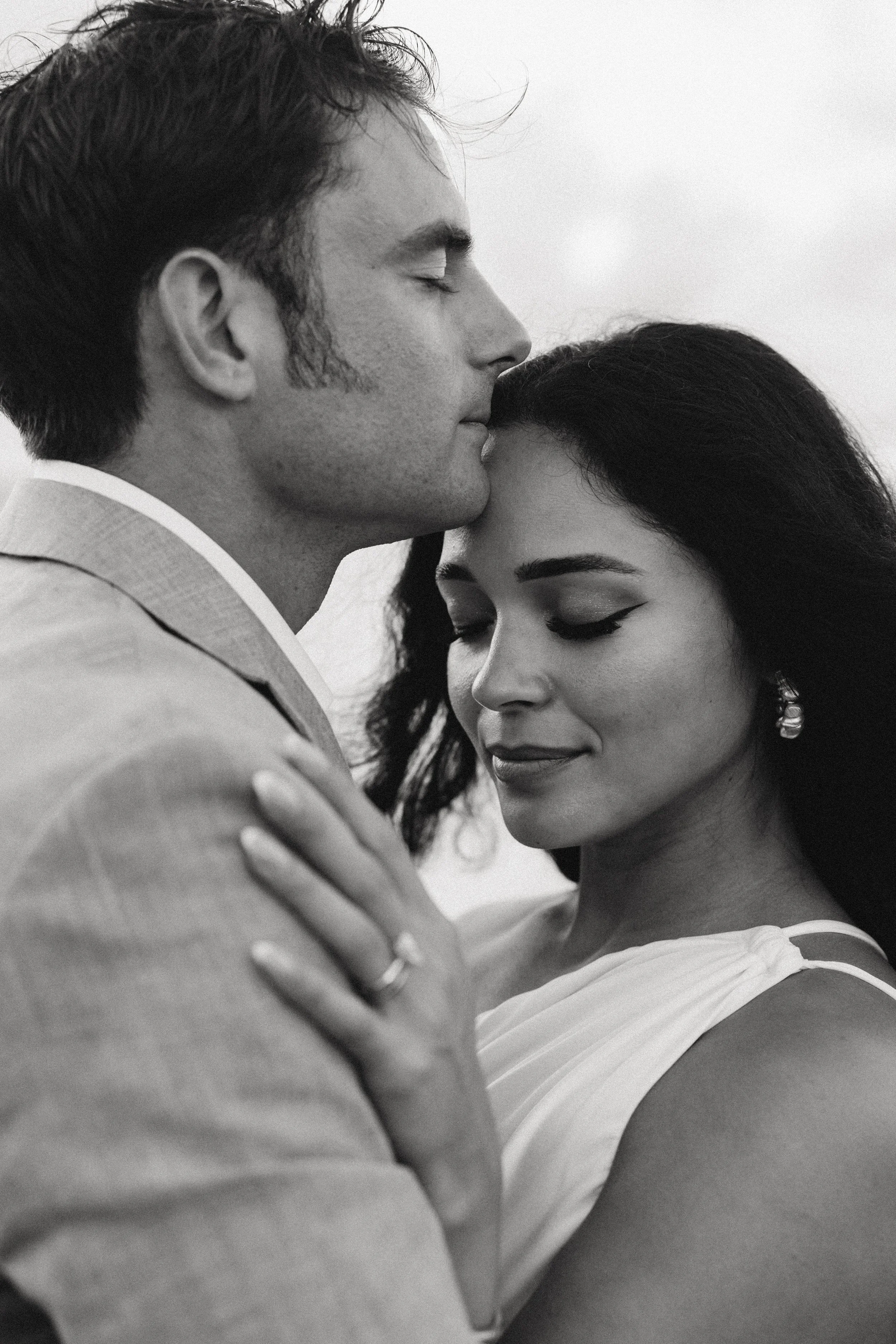 A black-and-white photo of a man and woman in a close embrace, with the man gently kissing the woman's forehead and the woman with her eyes closed, wearing earrings and an elegant dress.