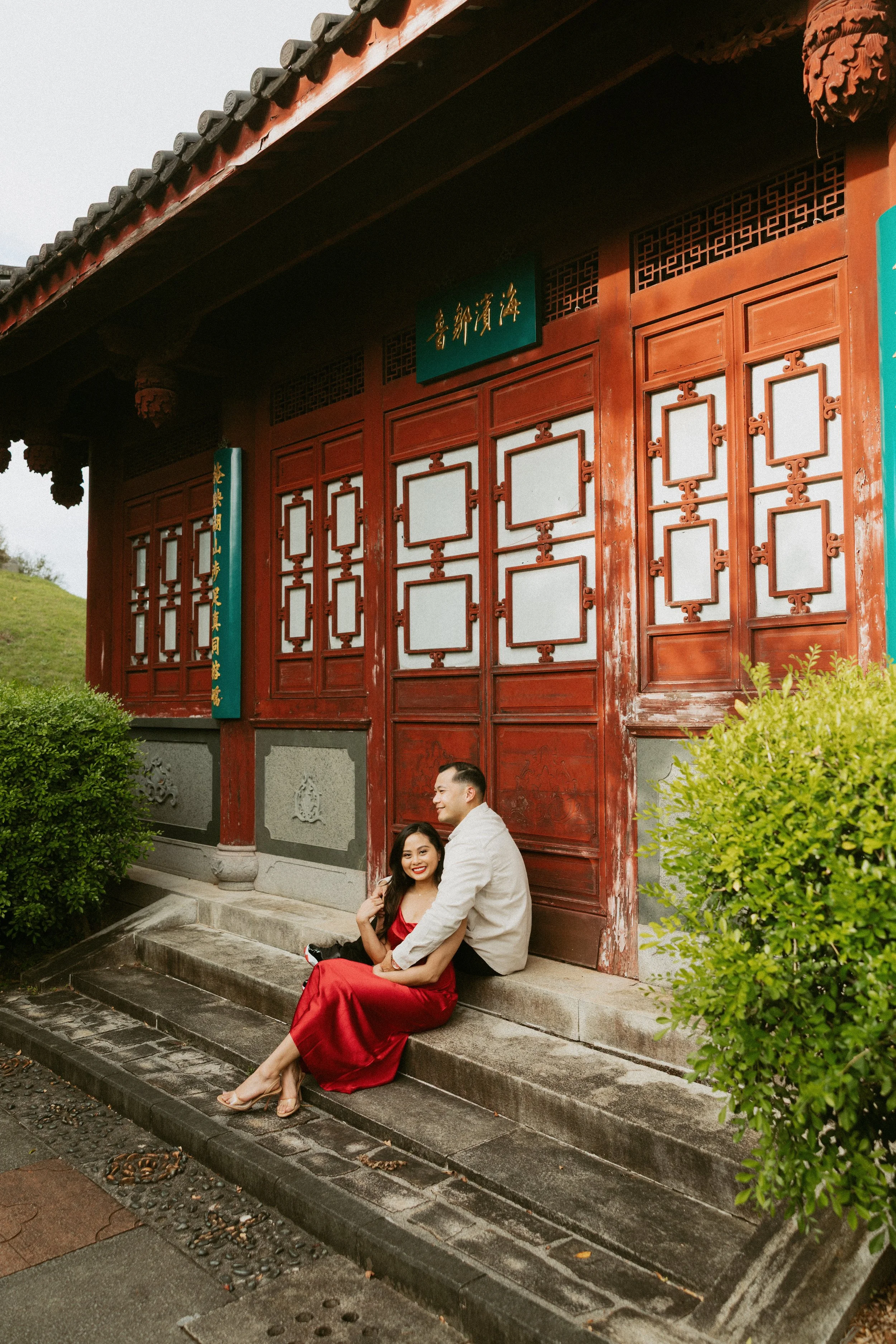 A couple sitting on stone steps outside a traditional Asian building with red wooden doors and decorative panels, surrounded by green shrubs.