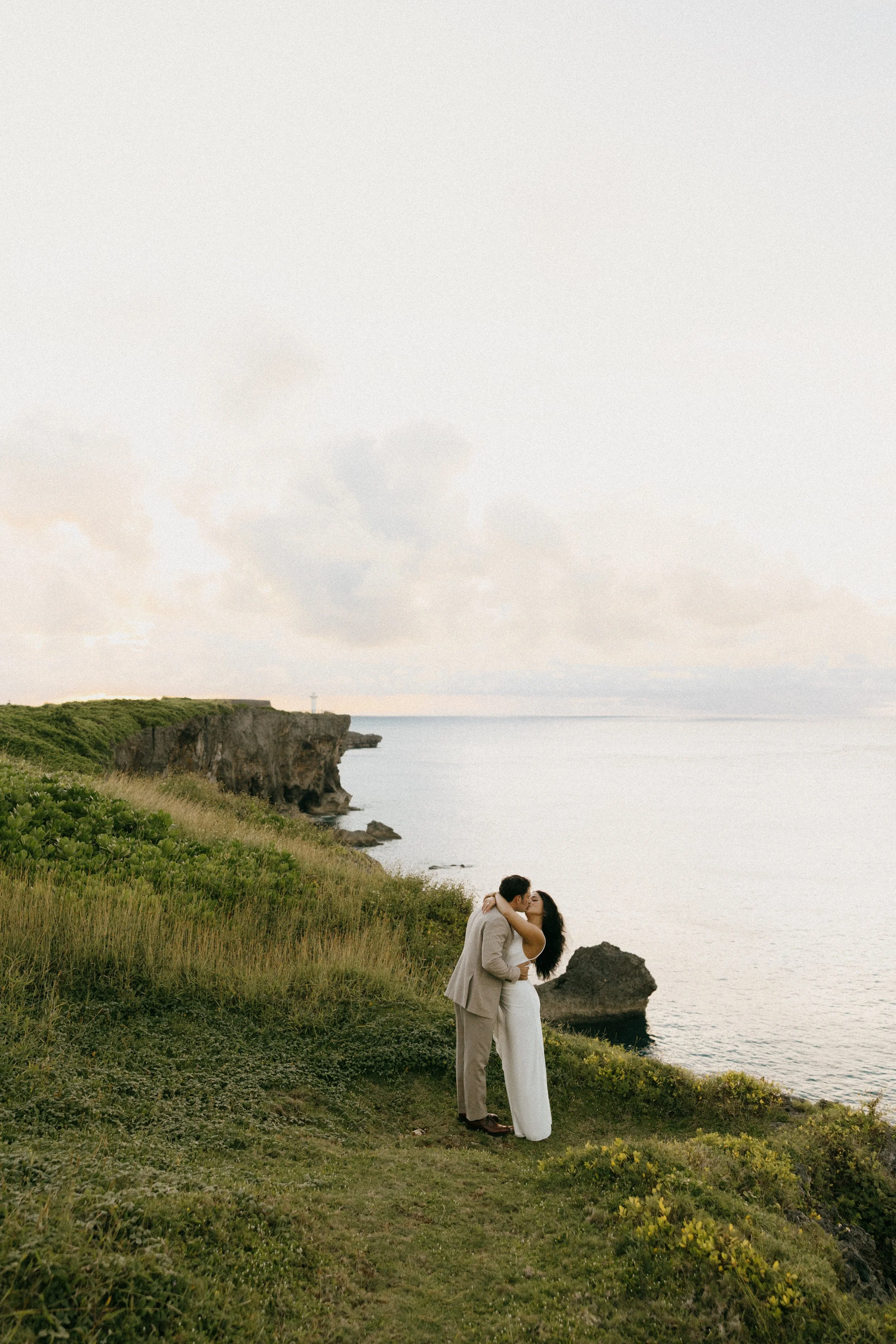 A couple dressed in wedding attire is kissing on a grassy cliff overlooking the ocean during sunset.