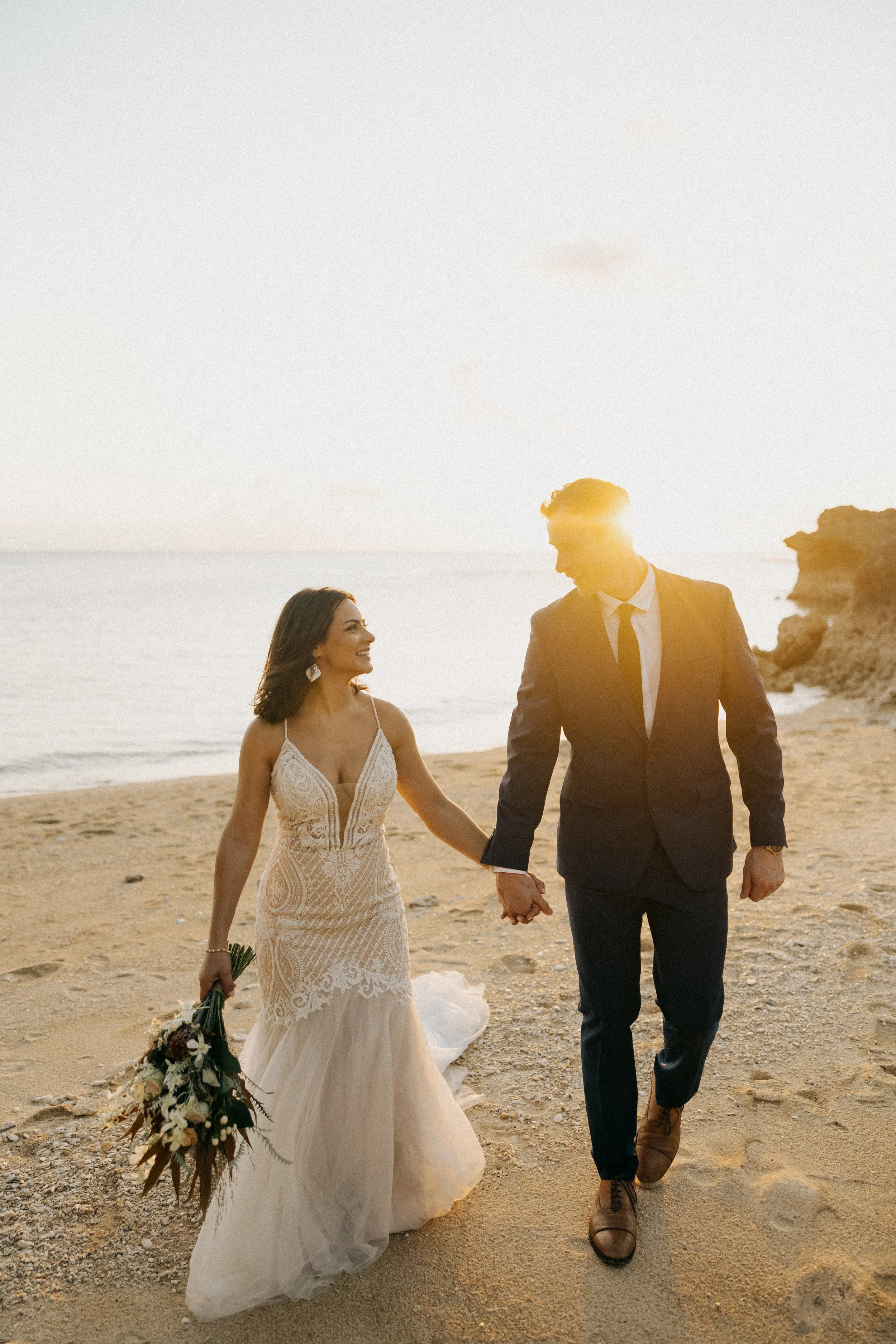 A bride and groom walking hand in hand on a beach at sunset, smiling and looking at each other, with the bride holding a bouquet.