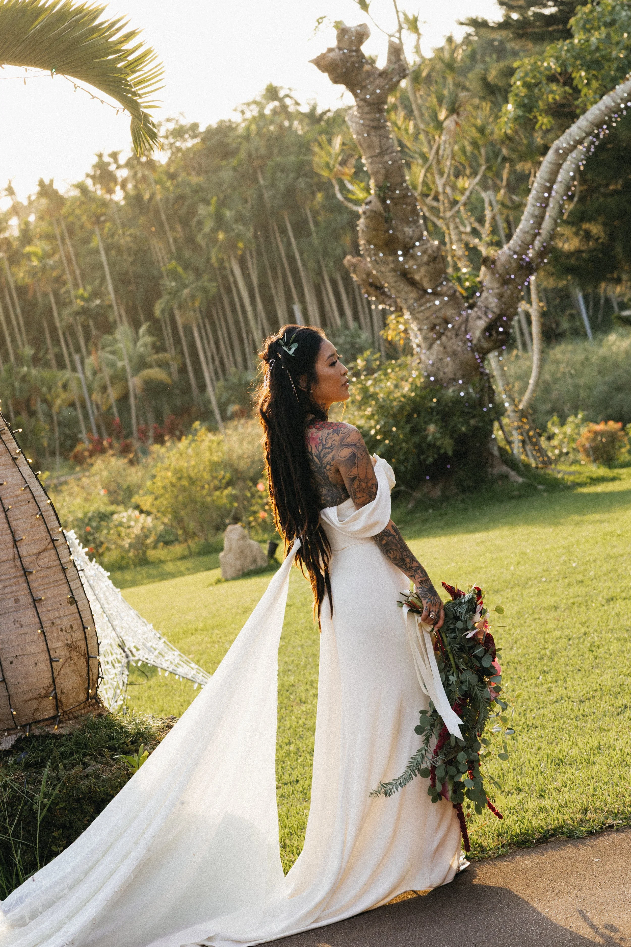 A bride in a white wedding gown holding a bouquet of flowers standing outdoors near a twisted tree decorated with string lights during sunset.