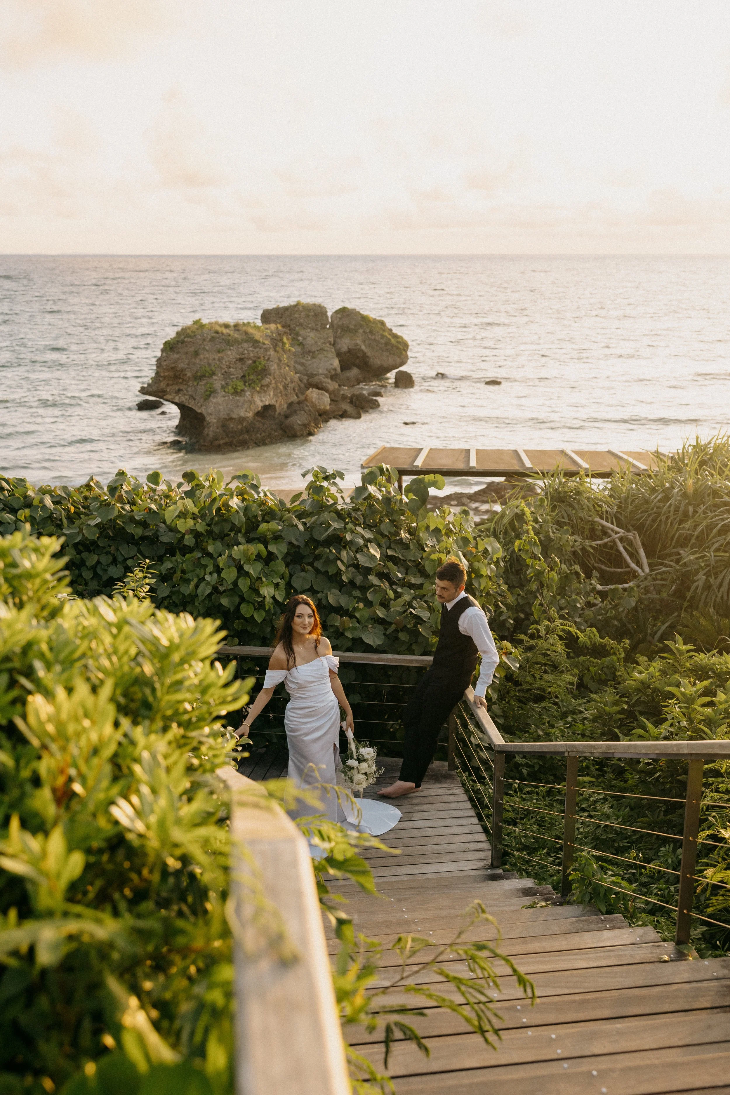 A couple dressed in wedding attire standing on a wooden pathway surrounded by green foliage with the ocean and large rocks in the background during sunset.